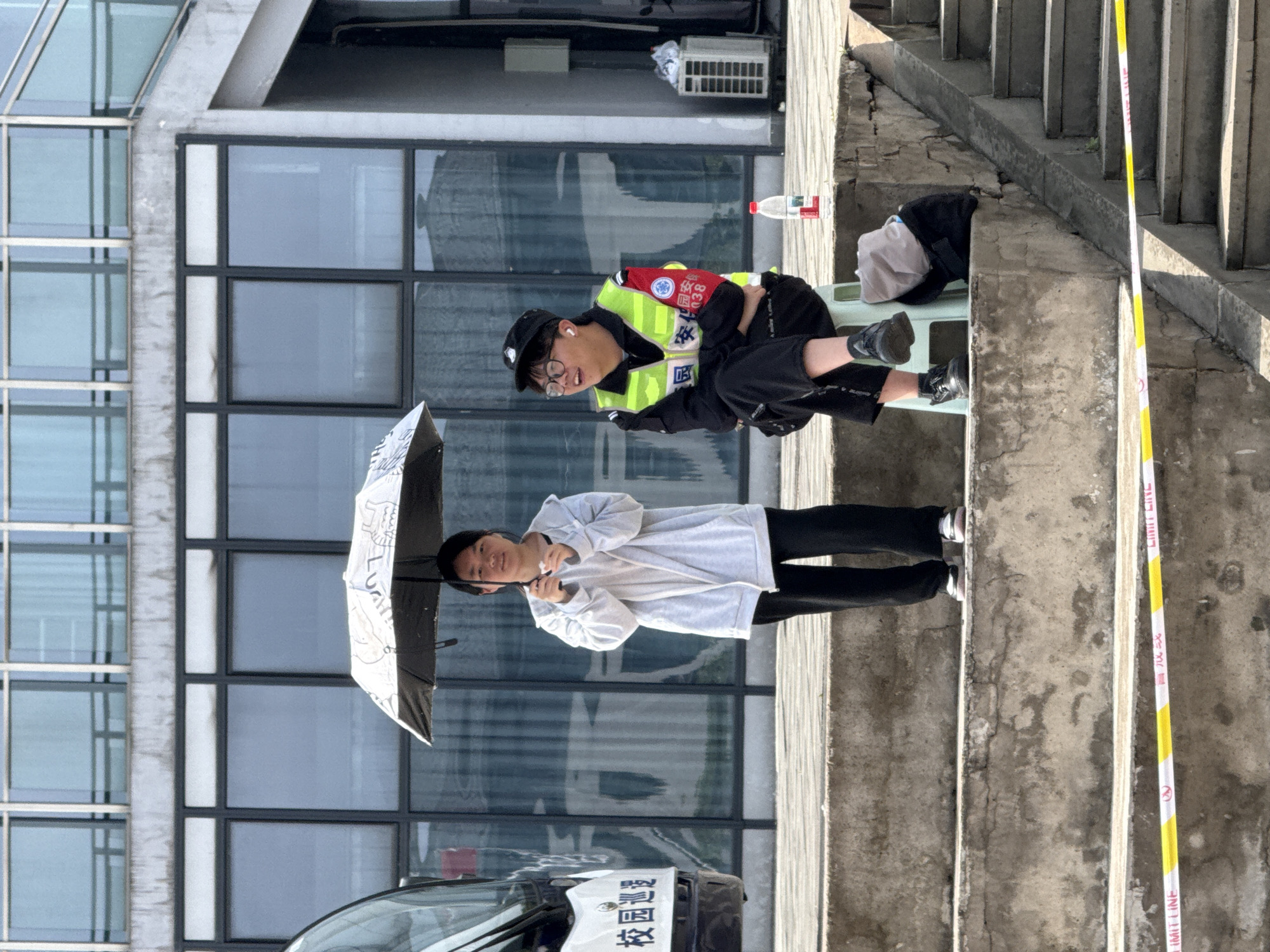 The image shows two people on a staircase outside a building. A person wearing a light grey hoodie and black pants holds a black umbrella and stands on the lower steps. Above and to the right, a person wearing a black cap, glasses, and a fluorescent yellow vest is seated on a small plastic chair. The scene is set against a modern building with large, multi-pane windows. The stairway is marked with yellow and white "LIMIT LINE" tape. The location is Jiangjin, China, inferred from the Chinese characters visible on the vest and a vehicle in the background.