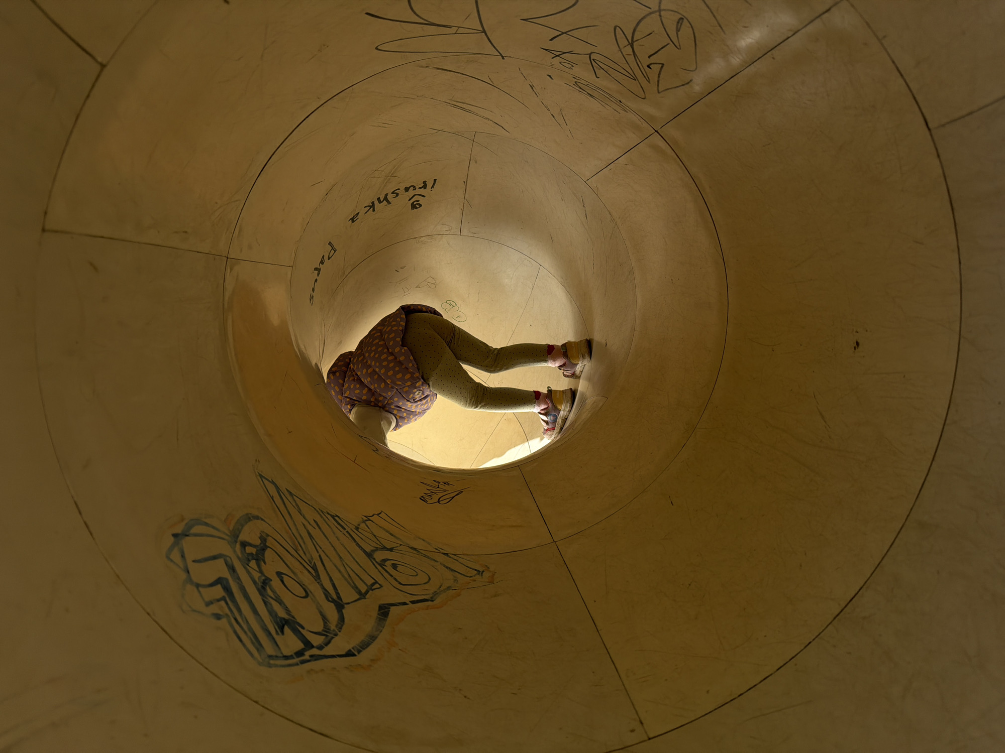 A child is partially visible inside a large, beige, circular tunnel. The tunnel has graffiti, including the name "Irushka Parus". The child is wearing a spotted jacket, light green pants, and colorful shoes. They appear to be crawling or standing within the tunnel. The location is Malakhovka, Russia.