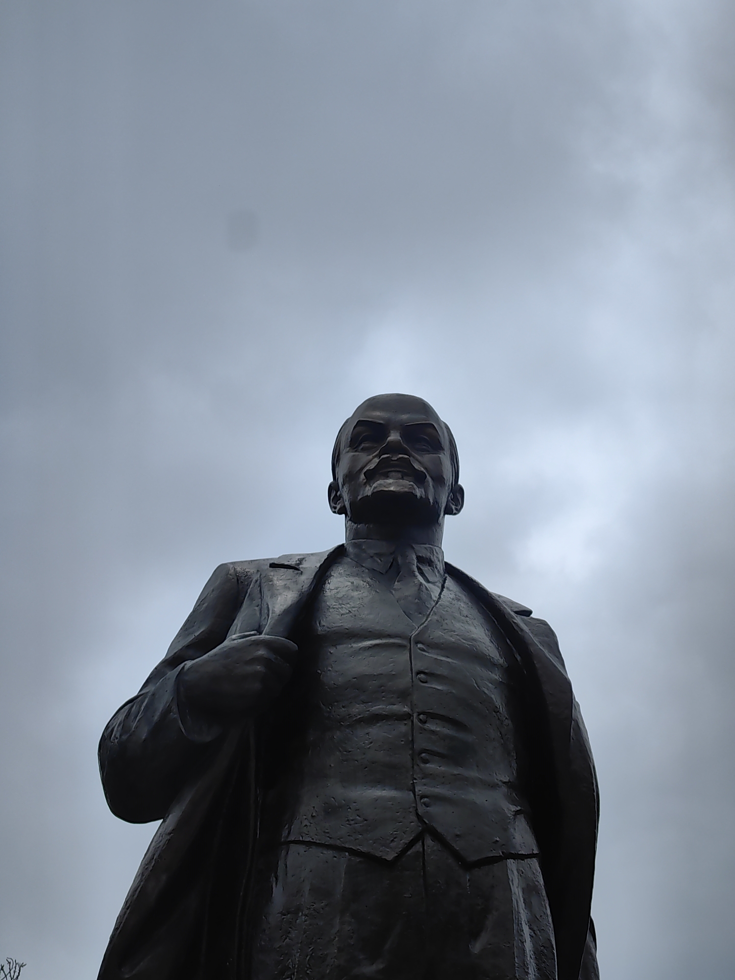 A low-angle shot depicts a statue of Vladimir Lenin against a cloudy sky. The bronze statue features Lenin in a suit, vest, and tie, with a coat draped over his shoulders. The location of the statue is in Hà Nội, Vietnam.