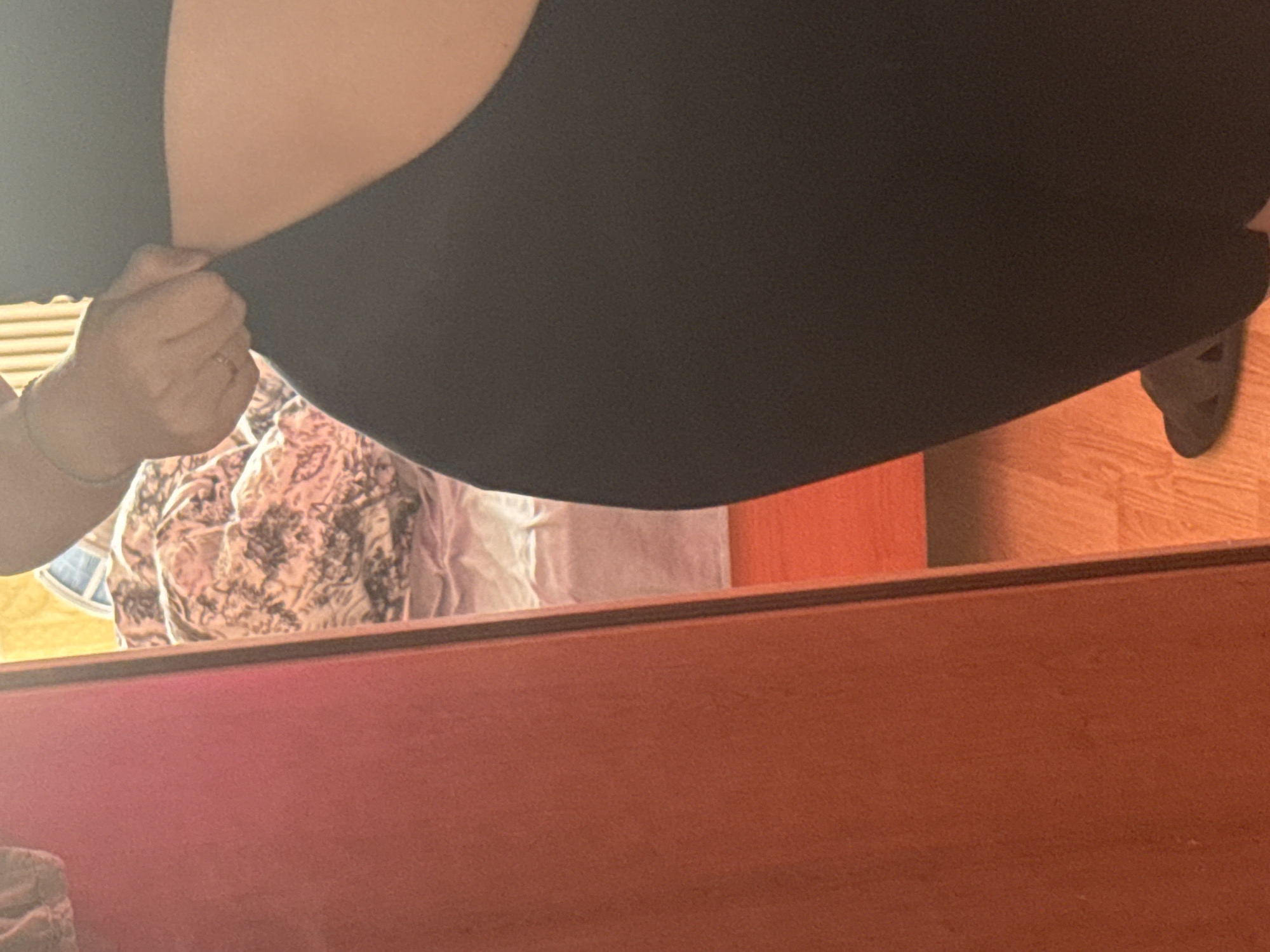 A person is standing in what appears to be a bedroom. The individual is wearing dark leggings and is pulling at the waistband. A large red piece of furniture, possibly a closet door or dresser, is visible on the left side of the frame. In the reflection of this furniture, a bed with patterned bedding and a portion of a window are visible. The floor is made of wood. A dark colored sandal is visible on the floor near the person's feet.