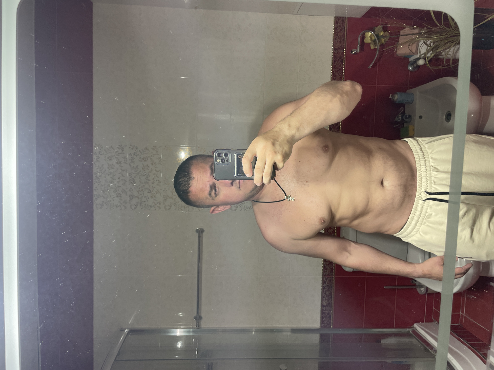A shirtless man with short dark hair and a cross necklace is taking a mirror selfie in a bathroom. The bathroom has tiled walls, a bidet, and a shower. The location is likely in Sochi, Russia.