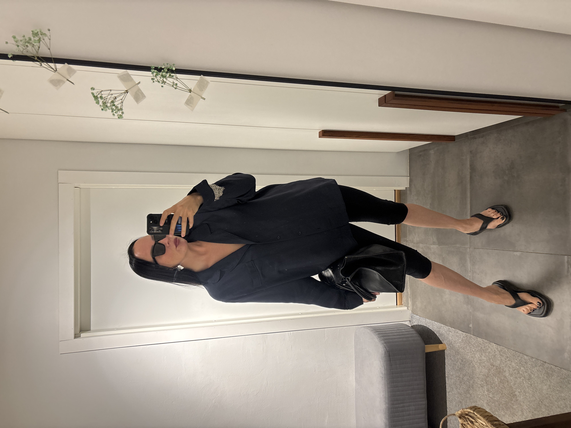 The image shows a person taking a selfie in what appears to be a retail dressing room or personal closet. The person is wearing black leggings, a black blazer or jacket, and black flip-flops. They are holding a smartphone in their hand, capturing their reflection. A black shoulder bag is also visible.
The environment features light-colored walls with decorative floral accents along the top of one wall. A gray, padded bench is located near the bottom of the frame. The floor is tiled in a gray concrete-like material. Based on the surroundings and the lack of distinctive landmarks, it is difficult to definitively pinpoint the exact location within Chaikovsky, Russia, without further context or identifying features. The space seems clean and well-maintained.