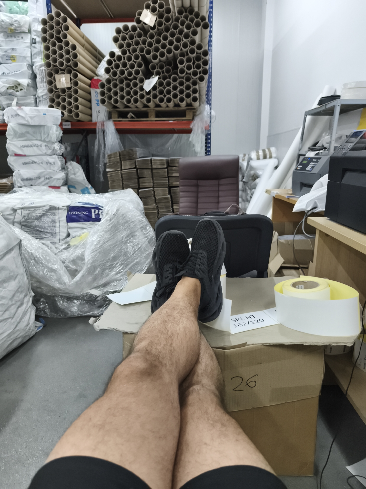 The image shows a first-person perspective of a person's legs and feet resting on a desk in a warehouse. The person is wearing black athletic shoes and black shorts. Their legs are crossed at the knee. The desk is cluttered with various items, including stacks of white paper, a roll of yellow tape, a printer, and some cardboard boxes. In the background, there are shelves stacked with rolls of cardboard tubes, large plastic-wrapped bags of material, and other industrial supplies. The overall environment appears to be a storage or distribution area within a warehouse.