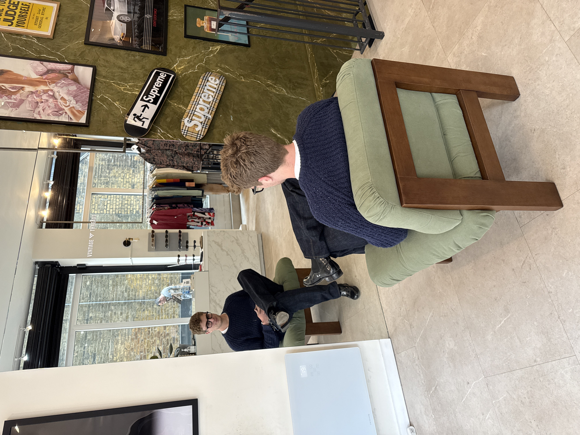Here is a description of the image:
The image captures an indoor scene, possibly a retail space, featuring two men seated in chairs. The man in the foreground is positioned with his back to the viewer and is partially obscured by an armchair. The second man, seated further back, is visible in a mirror reflection, creating a double visual effect. Both men are wearing dark-colored sweaters and dark jeans. The location appears to be a store, as there are clothing racks in the background and framed prints and decorative skateboards on the wall. The mirror reflects the interior of the store, revealing clothing displays and daylight streaming through the windows. The floor is tiled in a light beige color, adding to the store's modern aesthetic. The store sign reads Vintage Threads, indicating it may be a vintage or thrift store. The skateboards on the wall read Supreme. I have no information regarding location in London, United Kingdom.