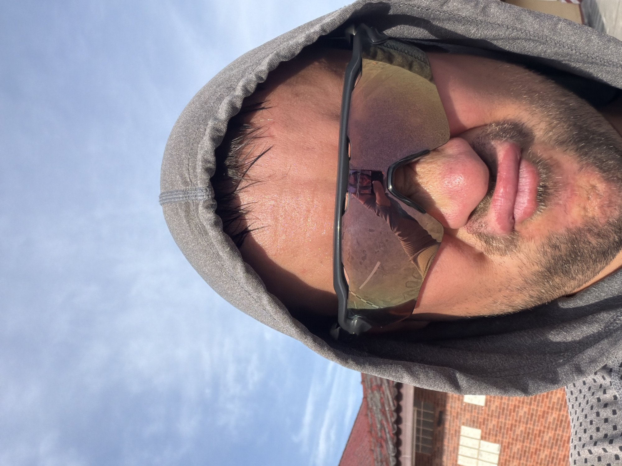A close-up, downward-angled, outdoor selfie shows a person's face partially obscured by a grey hood and tinted sunglasses. The person is making a puckered facial expression. The background includes a clear blue sky with some wispy clouds, a red-tiled roof of a building, and part of a brick wall. The scene is not located in Baan Mae Nam, Thailand; it appears to be a residential area.