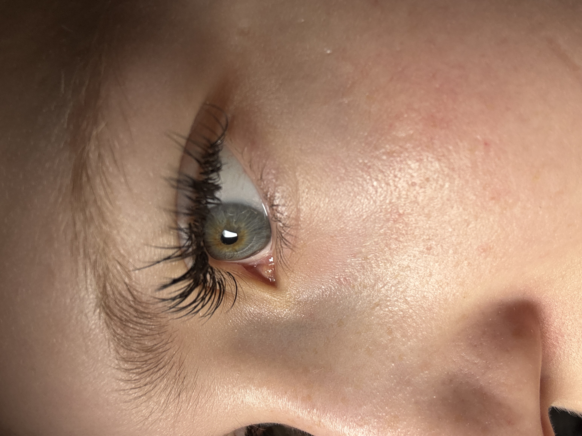 The image shows a close-up of a human eye and surrounding facial skin. The eye is light gray-green with dark eyelashes. The skin exhibits pores and minor imperfections, suggesting a youthful complexion. The eyebrow is light brown. No information is available to determine whether this image was taken in Chaikovsky, Russia, or any other location.