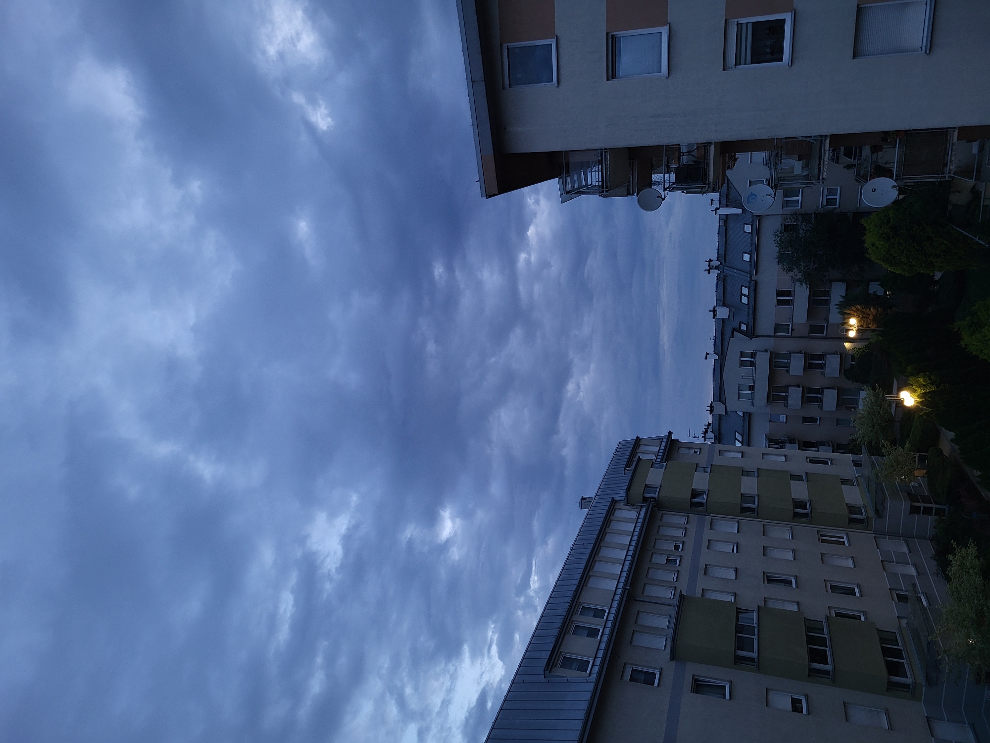 The image shows a view of apartment buildings against a cloudy sky. The buildings are modern and appear to be multi-story residential structures. The sky is overcast with dark, textured clouds, suggesting it might be evening or a storm is approaching. The lighting is dim, indicating a lack of direct sunlight. There are no people visible in the image. The foreground features the sides and balconies of the buildings, with some satellite dishes visible on one of the structures. Trees and greenery can be seen at the base of the buildings.