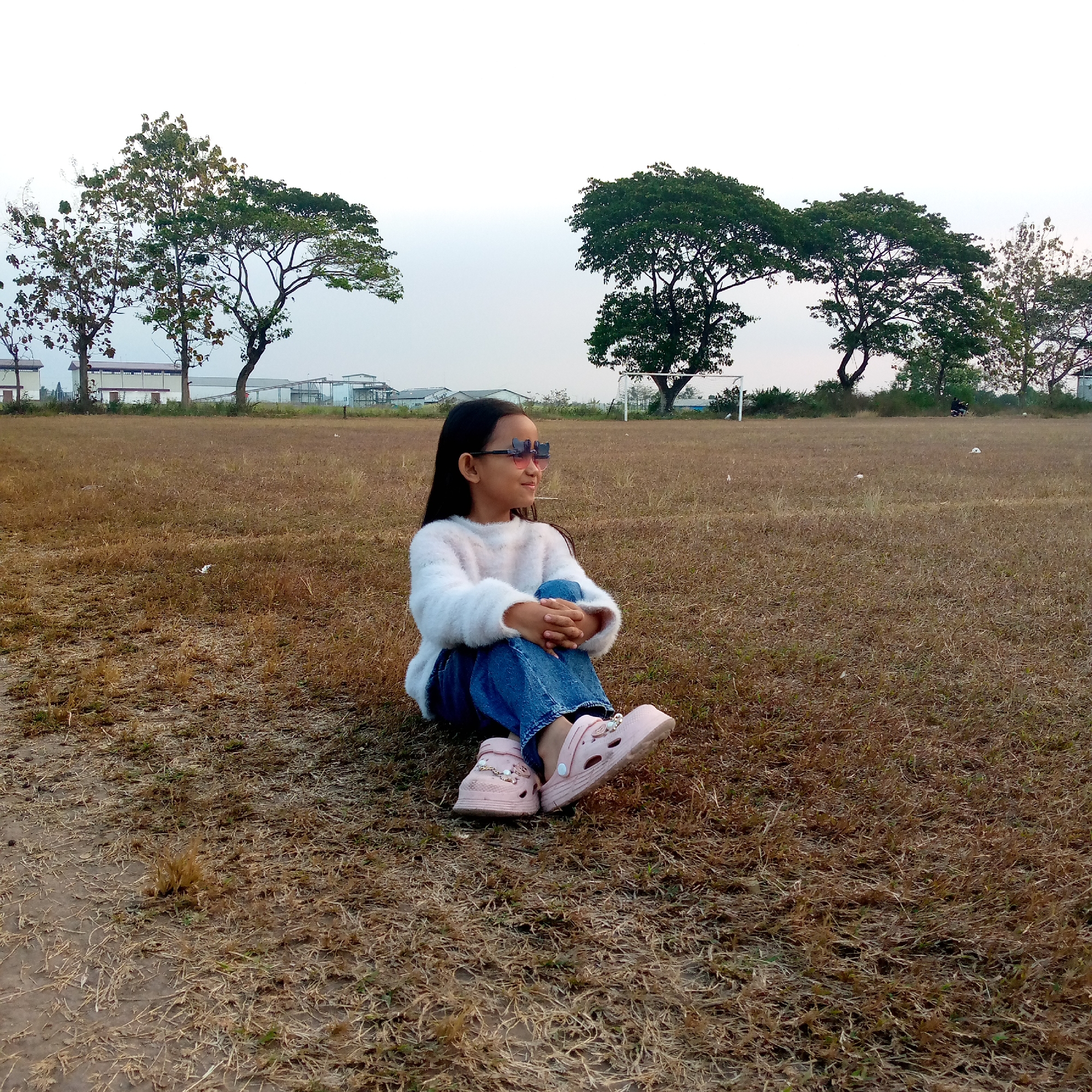 A young girl with long dark hair and pink butterfly-shaped sunglasses sits cross-legged on a field of dry, brown grass. She is wearing a fluffy white sweater and blue jeans. Her feet are adorned with pink clogs, decorated with pearl-like accents. In the background, several large trees with green foliage stand silhouetted against a pale sky. To the left, industrial buildings with white walls and red roofs are visible. A white soccer goal is situated on the right side of the frame. The overall setting appears to be an open field, possibly on the outskirts of the city of Pacing, Indonesia, indicated by the presence of industrial structures and a soccer goal.