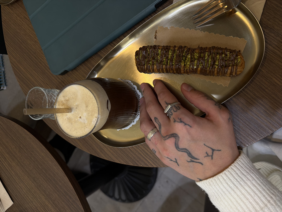 An overhead shot captures a casual dining or coffee break scene featuring a person's tattooed hand, a drink, and a dessert on a golden tray.
In the foreground, a left hand rests palm down on a shiny, oval-shaped golden or brass-colored tray. The hand is notably tattooed with a stylized snake design winding across the back of the hand, and several smaller, abstract, twig-like markings on the fingers and knuckle areas. Two rings adorn the fingers: a textured band on the ring finger and a geometric, possibly hexagonal, ring on the middle finger. The wrist is partially visible, showing the cuff of a light-colored, possibly off-white, ribbed long-sleeved sweater.
On the golden tray, to the left of the hand, sits a tall, ribbed glass holding a dark beverage with a thick layer of light brown foam on top, suggesting an iced coffee or similar espresso-based drink. A light-colored, possibly bamboo or paper, straw is inserted into the drink. The glass rests on a small, white, decorative doily. Another empty, clear glass is partially visible just to the left of the main drink.
To the right of the hand, also on the golden tray, is a dessert that resembles an eclair or a churro. It's elongated and cylindrical, covered generously with dark chocolate drizzle and sprinkled with green crushed nuts, likely pistachios. The dessert rests on a piece of light brown parchment paper. A silver fork is visible at the top right of the tray, also resting on the parchment paper.
The objects are placed on a dark wooden table with a visible grain. In the upper left corner of the image, part of a teal or turquoise-colored tablet or notebook cover is visible, suggesting other items are on the table. The overall scene conveys a relaxed, indoor setting, likely a cafe or restaurant. The lighting is soft and even, indicating a well-lit environment. There is no visible text in the image.
