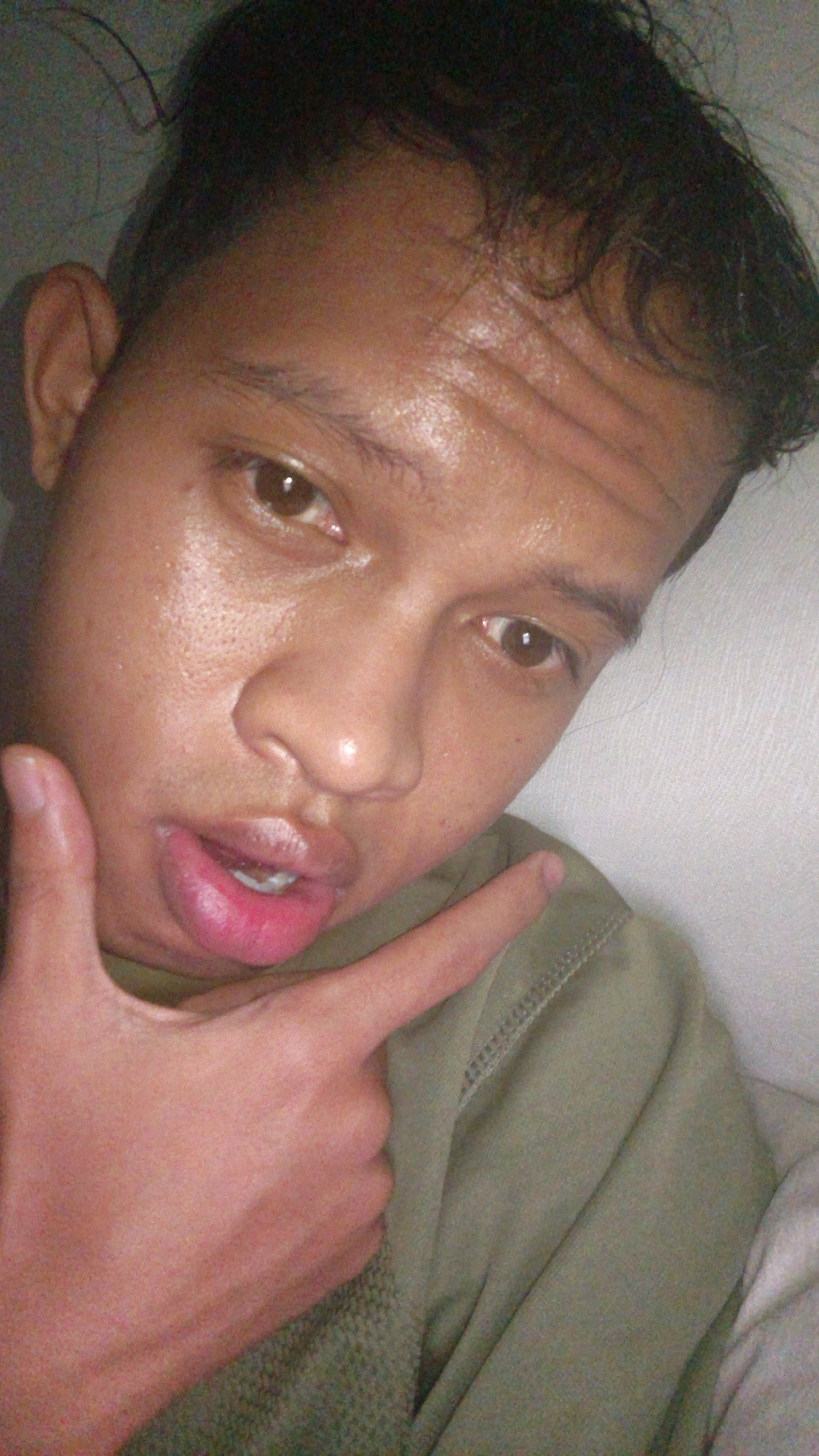 A close-up, head-and-shoulders selfie shows a person with dark hair and brown eyes, looking directly at the camera. The person's skin appears to be shiny with sweat. They are wearing a green shirt. The person's right hand is raised, with the index finger pointing towards their own face, near their mouth. The lips are slightly parted, revealing white teeth. The background is a plain, light-colored wall. The image was taken indoors. There is no information available regarding the specific location within Pundungsari, Indonesia.