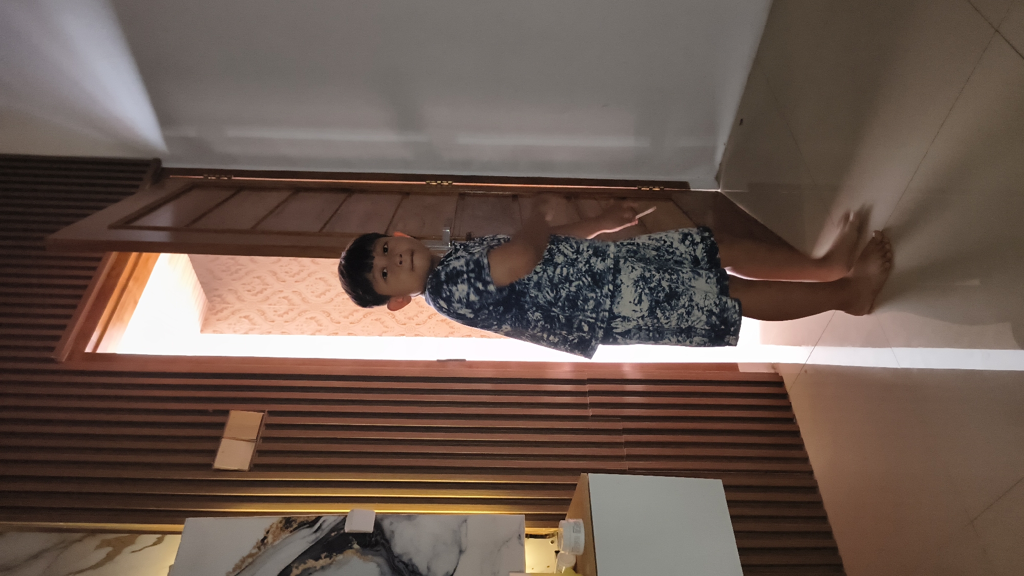 The image shows a young boy standing in a doorway, looking towards the camera. He is wearing a dark blue and white patterned shirt and shorts. His skin is tan, and he has dark hair. The boy's left arm is slightly bent, and he appears to be holding something thin and white in his hand, though it is mostly obscured. His feet are bare, and he is standing on a light-colored tiled floor.
Behind the boy, the doorway opens into a room with patterned wallpaper and a bright light source, possibly a window or a light fixture. The doorway itself is made of dark wood, with horizontal slatted wood paneling on the wall to the left. The overall lighting suggests it might be daytime, with natural light coming from the room beyond the doorway and some artificial light illuminating the area. The scene appears to be indoors, likely in a home, given the interior design elements like the wooden paneling and tiled floor. The boy has a neutral expression. There is no visible text in the image. The orientation of the image is on its side, with the top of the boy's head appearing on the left side of the frame.