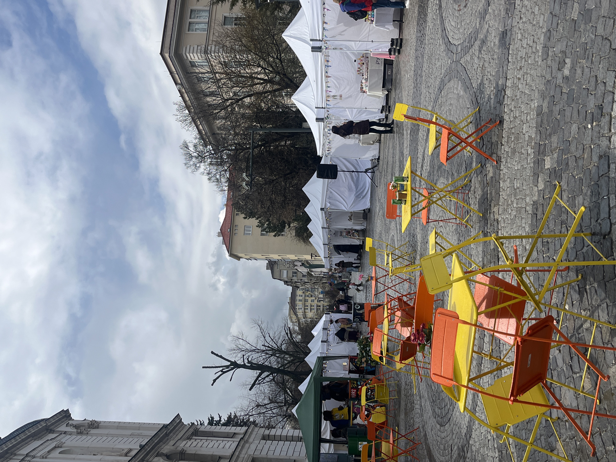 The image shows a street market scene in Sofia, Bulgaria. White vendor tents are lined up along a cobblestone street, and there are yellow and orange chairs and tables scattered around in the foreground. People are walking around the market, and some are looking at the merchandise being sold. Buildings are visible in the background, and the sky is cloudy.