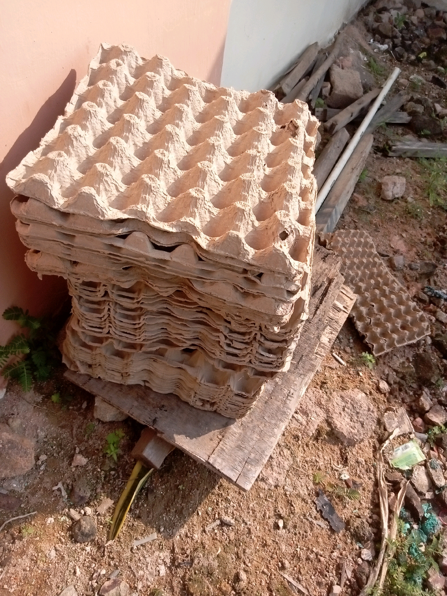 A stack of used, beige cardboard egg cartons is piled on a wooden board. The cartons are made of molded pulp and have an uneven, textured surface with individual indentations for eggs. The stack is positioned outdoors, leaning against a light-colored wall on the left side of the frame. The ground beneath and around the cartons is composed of dry soil, gravel, and debris, including scattered rocks, broken pieces of concrete, and dried vegetation. To the right of the egg cartons, a white PVC pipe and several pieces of weathered wood are visible, lying on the ground among the rocks and dirt. The scene appears to be an outdoor storage area or a waste disposal site, possibly in a less maintained part of a residential or industrial area in Batam, Indonesia. The lighting suggests it is daytime, with shadows cast by the wall and the egg cartons.