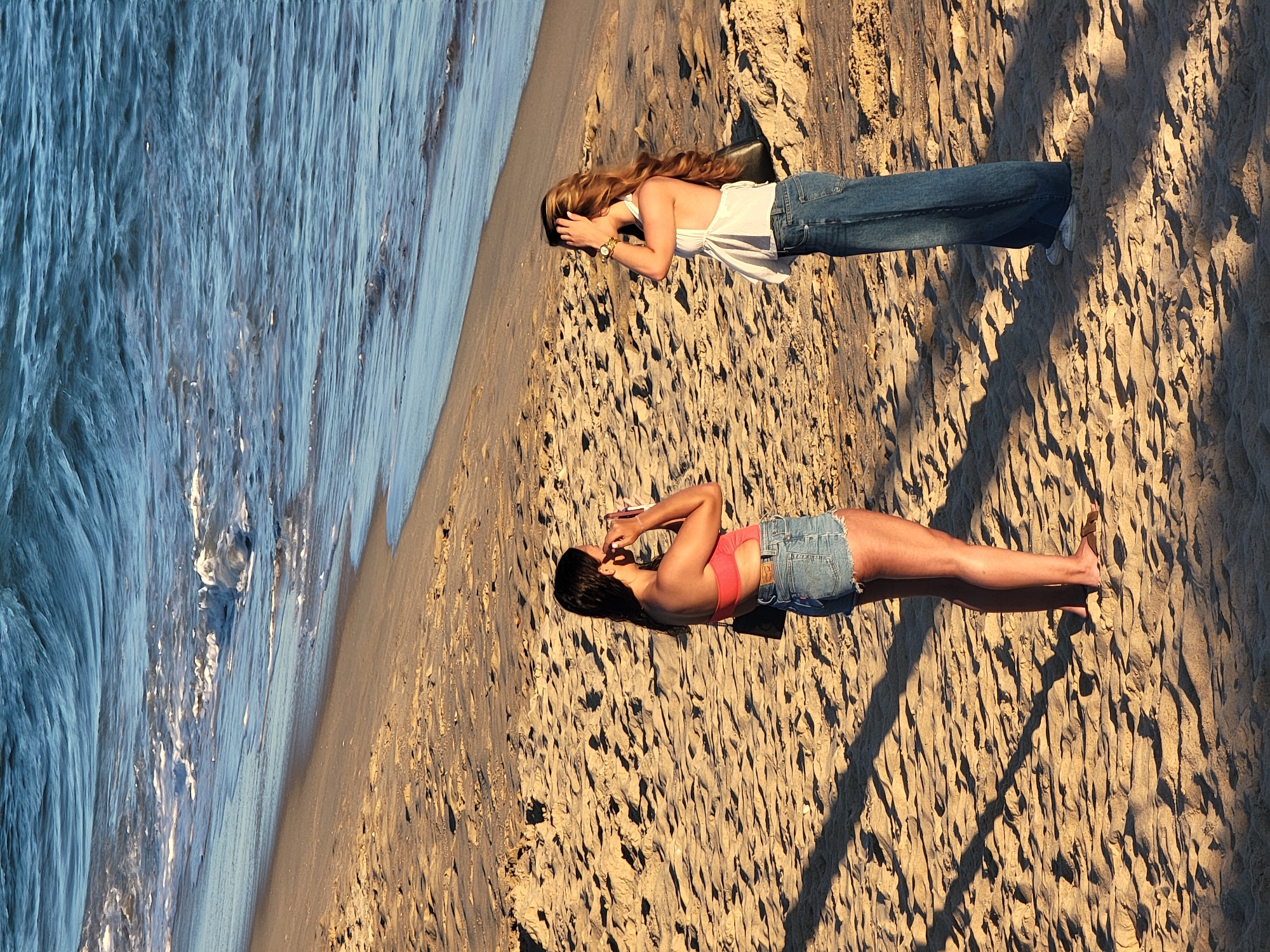 Two women stand on a sandy beach in Quarteira, Portugal, near the ocean. The woman on the left wears a coral-colored bandeau bikini top and denim shorts, holding a pink object to her face as if taking a picture. She has dark hair and is barefoot. The woman on the right has long, wavy blonde hair and wears a white halter top and loose-fitting blue jeans. She has her hand up to her head, adjusting her hair or shielding her eyes. The beach is covered in fine sand with footprints and shadows cast by an unseen light source. The ocean, with gentle waves and white foam, is visible in the background.