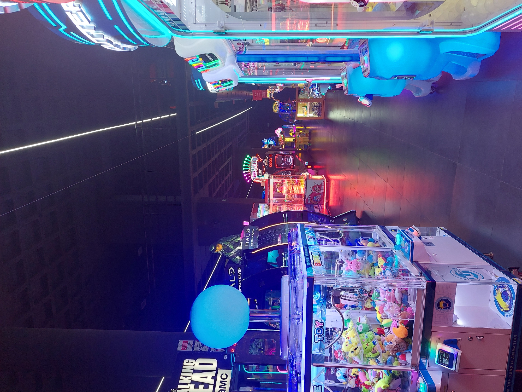 This image captures the vibrant interior of a modern arcade or entertainment center, likely in Buenos Aires, Argentina. The scene is characterized by a long, dimly lit aisle lined with numerous brightly illuminated arcade games, creating a dynamic and engaging atmosphere.
In the foreground and extending into the midground, various types of arcade machines are visible. Several claw machines, filled with colorful plush toys, stand prominently. To the right, blue race car-shaped seats indicate a driving simulation game. Further down the aisle, a "Halo: Fireteam Raven" game cabinet, featuring a detailed character graphic, is clearly identifiable. Another game, "The Walking Dead AMC," is visible on a cabinet in the foreground, near a large, glowing blue spherical light installation. Other games with signs like "GOOD TI" and "FREE LUCK" contribute to the diverse array of entertainment options. A small red sign further down the aisle, partially obscured, appears to say "CAJA," which is Spanish for "cashier," reinforcing the location context.
The setting is an indoor space, probably in the evening or at night, given the reliance on artificial lighting from the games and neon strips. Long, horizontal white lights are integrated into the ceiling structure, adding to the contemporary design. The floor is dark and tiled. No people are visible, but the arrangement of machines suggests an environment designed for interactive play and entertainment. The overall mood is one of excitement and fun, primarily conveyed through the dazzling array of colored lights emanating from the games. Visible text includes "THE WALKING DEAD AMC," "HALO FIRETEAM RAVEN," "TOY," "WE TOY TONG LI ANIMATION," "FREE LUCK," "GOOD TI," and "CAJA."