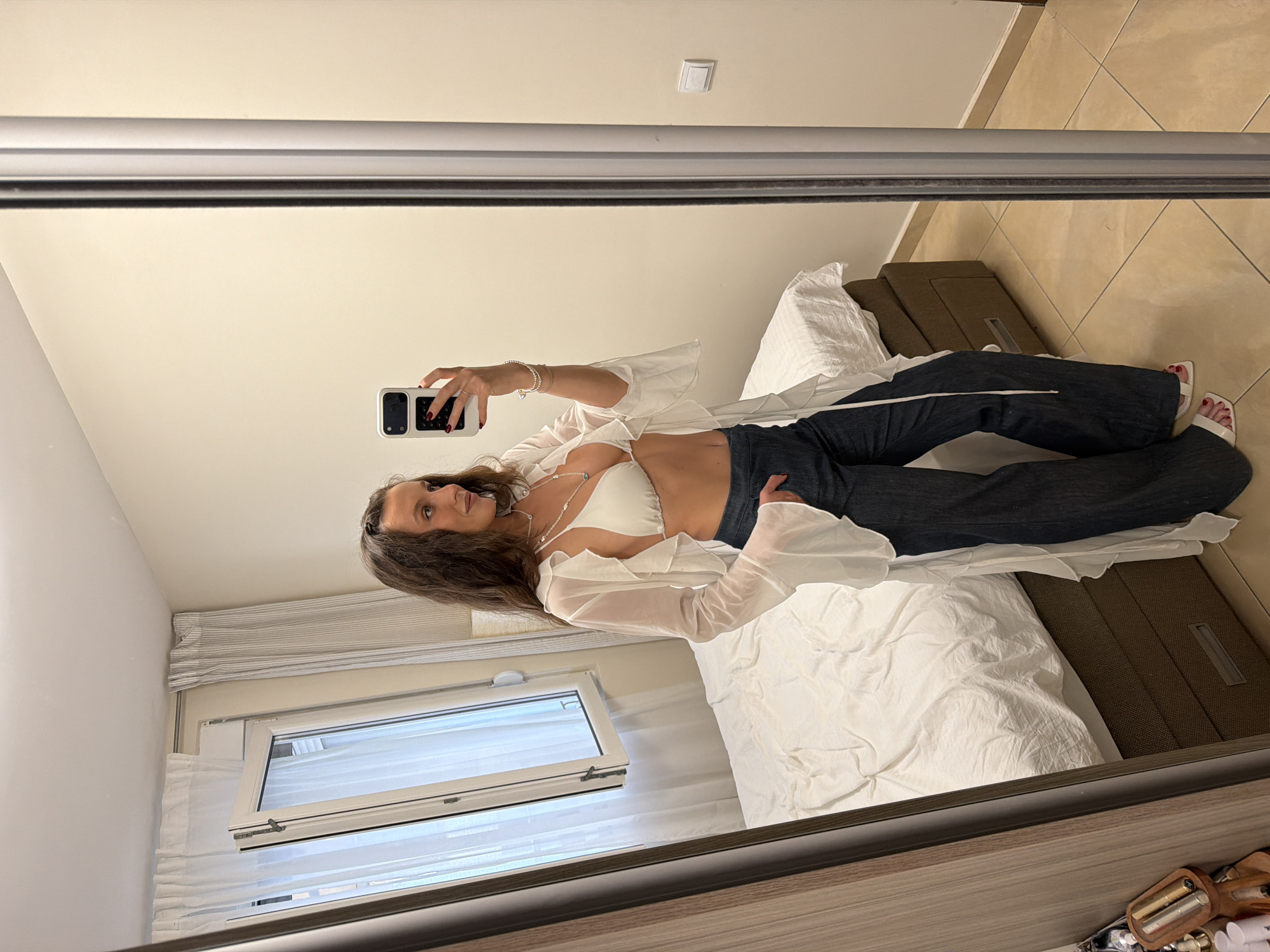 A woman is seen in a mirror selfie indoors. She is wearing a white bikini top with a sheer white long-sleeved shirt, dark jeans, and white heeled sandals. She is holding a smartphone in her right hand, presumably taking the photograph. Her hair is long and wavy, and she is wearing jewelry on her neck and wrist.
The room has a bed with white sheets and a brown headboard. A window with sheer curtains is visible in the background. The floor is tiled. The overall setting appears to be a bedroom, potentially in a hotel or apartment. It is not possible to verify that the location is in Budva, Montenegro from this image alone.