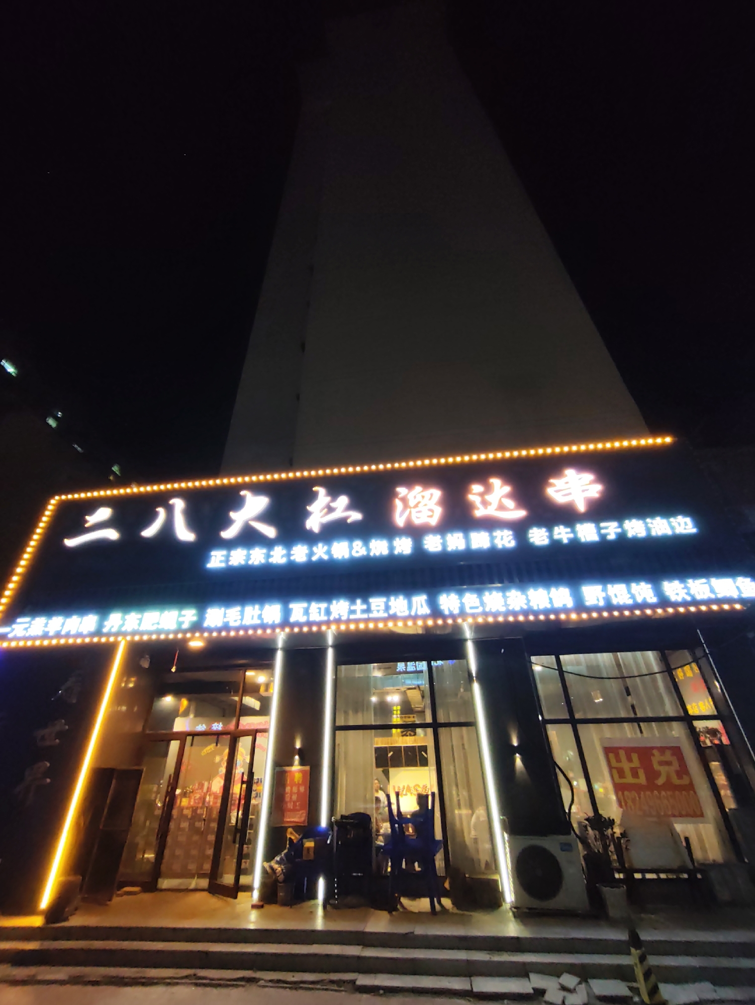 The image shows the exterior of a restaurant at night in Lindian, China. The restaurant's name, "二八大灶 溜达串," is prominently displayed on a lit-up sign. Beneath the main name, smaller text lists various dishes offered, including "正宗东北老火锅 & 烤," "一家串串," "烤肥肠," "溜达串," "爆肚," "烤豆皮," "特色烧杂," "野馄饨," and "铁板鲫鱼." The restaurant has large glass windows and a double door entrance. Inside, chairs are visible, and some people appear to be present. There is an air conditioning unit mounted on the exterior wall. The street in front of the restaurant is dark, with some steps leading up to the entrance. The overall impression is of a typical urban restaurant at night.