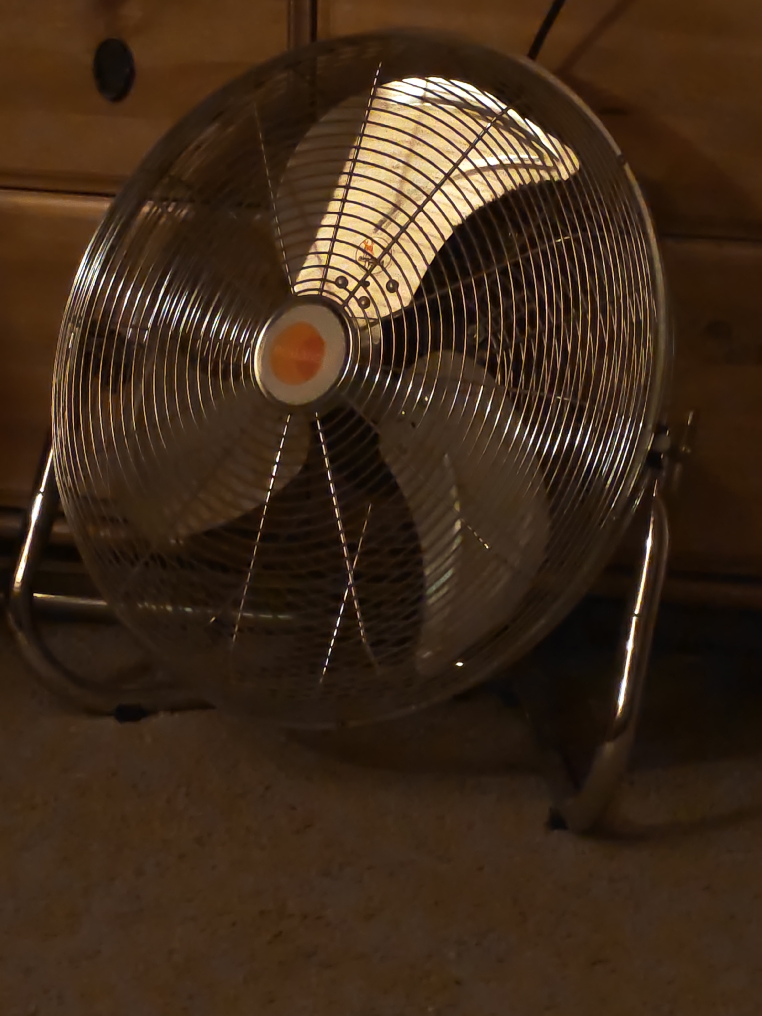 A metallic, oscillating fan is positioned on a carpeted floor, situated in front of a wooden dresser. The fan is turned on, as indicated by the light emanating from its center and the slight blur of the blades. The lighting in the room is dim, casting shadows. The scene is located in Novaya Polyana, Russia.