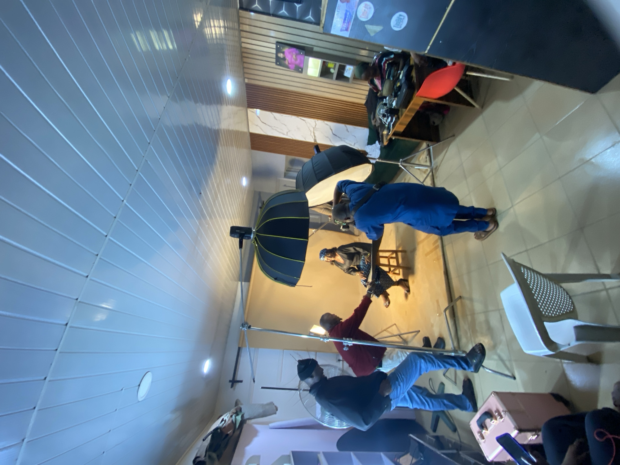 The image depicts an indoor photography session taking place in Bukuru, Nigeria. The scene is set in what appears to be a studio or a well-lit room with a tiled floor and a corrugated metal ceiling. Several people are present, engaged in various aspects of the photoshoot.
In the foreground, a person in a blue outfit is standing and appears to be directing or posing for the camera. Further back, another individual in a red jacket is actively involved, possibly adjusting equipment or interacting with the subject. A third person, wearing a dark hoodie and a beanie, is also visible. In the background, a woman is seated, possibly the main subject of the photoshoot.
The setup includes professional lighting equipment, such as large umbrella softboxes mounted on stands, which are typical for studio photography. There are also chairs and what looks like a portable vanity case or makeup kit, suggesting preparations for the shoot. The overall atmosphere appears to be focused and professional.
The time of day is not explicitly indicated, but the indoor lighting suggests it could be anytime. There is no indication of weather conditions. The location cues, such as the specific style of ceiling and flooring, along with the textual context identifying the location as Bukuru, Nigeria, provide geographical context. No specific emotions are overtly displayed, as the focus seems to be on the technical and practical aspects of the photography session. There are no discernible visible text elements in the image, apart from what might be on the items in the background shelves which are not legible.