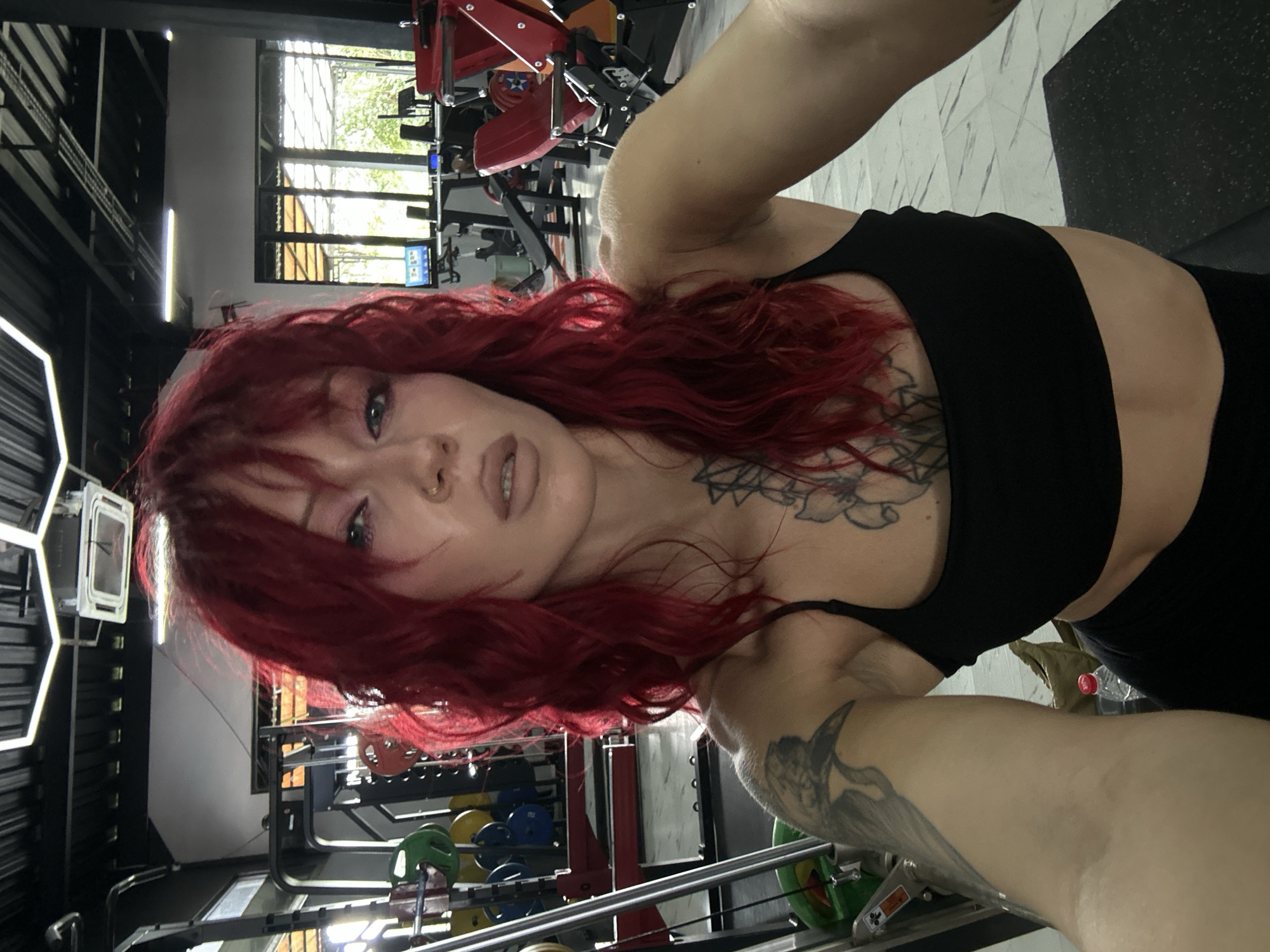 A woman with vibrant red hair is taking a selfie in a gym located in North Kuta, Indonesia. She is wearing a black sports bra and black leggings. Her hair is styled in loose waves, and she has dramatic eye makeup. She has tattoos on her left arm and chest. In the background, various weightlifting equipment, including weight plates and machines, are visible. Large windows allow natural light to enter the gym, illuminating the space.