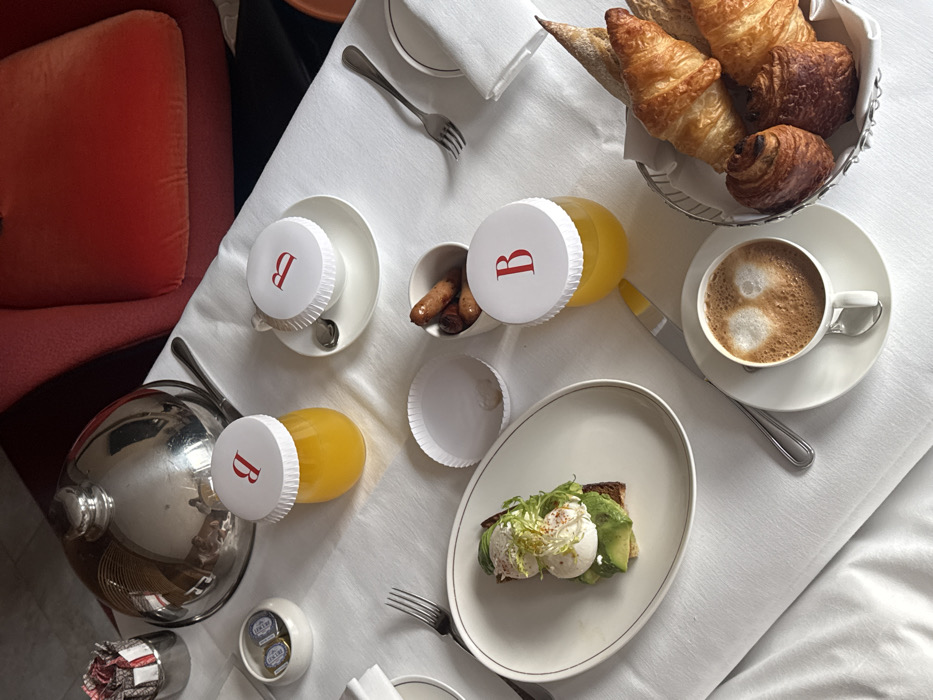 The scene depicts a meticulously arranged breakfast service laid out on a white tablecloth, likely within a high-end hotel room or private dining area. No individuals are present in the image.
The table prominently features an oval white plate holding avocado toast topped with two poached eggs and green garnish. To its left, a fork is positioned. A small white bowl contains several cooked sausages, and an empty white paper cup liner is also visible. Three items—two glasses containing orange juice and a white teacup on a saucer—are each covered with a white lid bearing a distinct red 'B' logo. A white cup of coffee, possibly a cappuccino, with latte art (two distinct foam hearts) is set on a saucer with a spoon, accompanied by a butter knife. A silver wire basket holds an assortment of baked goods, including multiple croissants and other pastries.
In the upper left quadrant, a silver domed cloche rests on a platter, with a serving utensil positioned beside it. A small white ceramic dish contains two individual packets of condiments, and a patterned napkin is neatly folded into a silver holder. The background reveals a section of a plush, reddish-orange upholstered armchair or sofa, indicating a comfortable, furnished indoor environment.