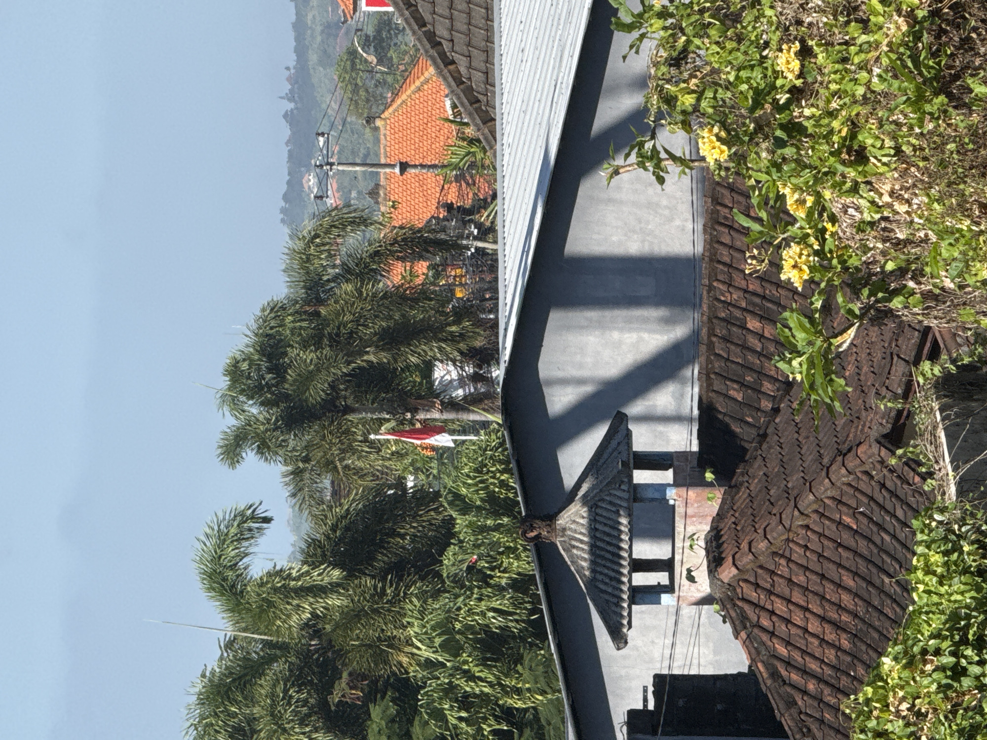 View of a residential neighborhood in Kuta, Indonesia, during daylight hours. The foreground contains buildings with tiled roofs, interspersed with dense green vegetation, including trees and flowering vines. In the background, more houses are visible, some with orange tile roofs. Utility poles with power lines can be seen extending across the scene. A glimpse of the sky is visible in the upper left corner. A small Indonesian flag is seen waving amidst the palm trees.