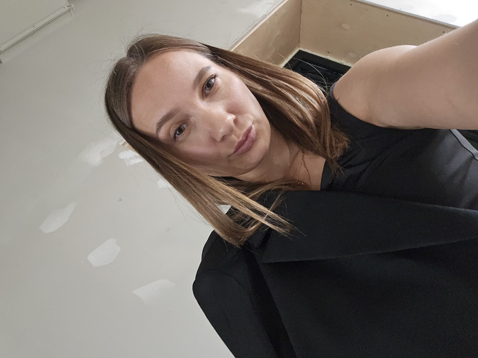 A close-up selfie captures a woman with long, straight brown hair, brown eyes, and fair skin, looking directly at the camera with a subtle pout. She is wearing a black top or jacket, with a faint necklace chain visible around her neck. Her right arm is partially visible in the upper right portion of the frame, indicating she is holding the camera to take the photograph. The background consists of light-colored, seemingly unfinished interior walls, with several white patches, possibly from spackle or primer. A wooden structure is visible in the upper right background, adjacent to a dark opening or recess. The overall impression is an indoor setting that may be under renovation. The photograph is situated in Chaikovsky, Russia. There is no visible text in the image.