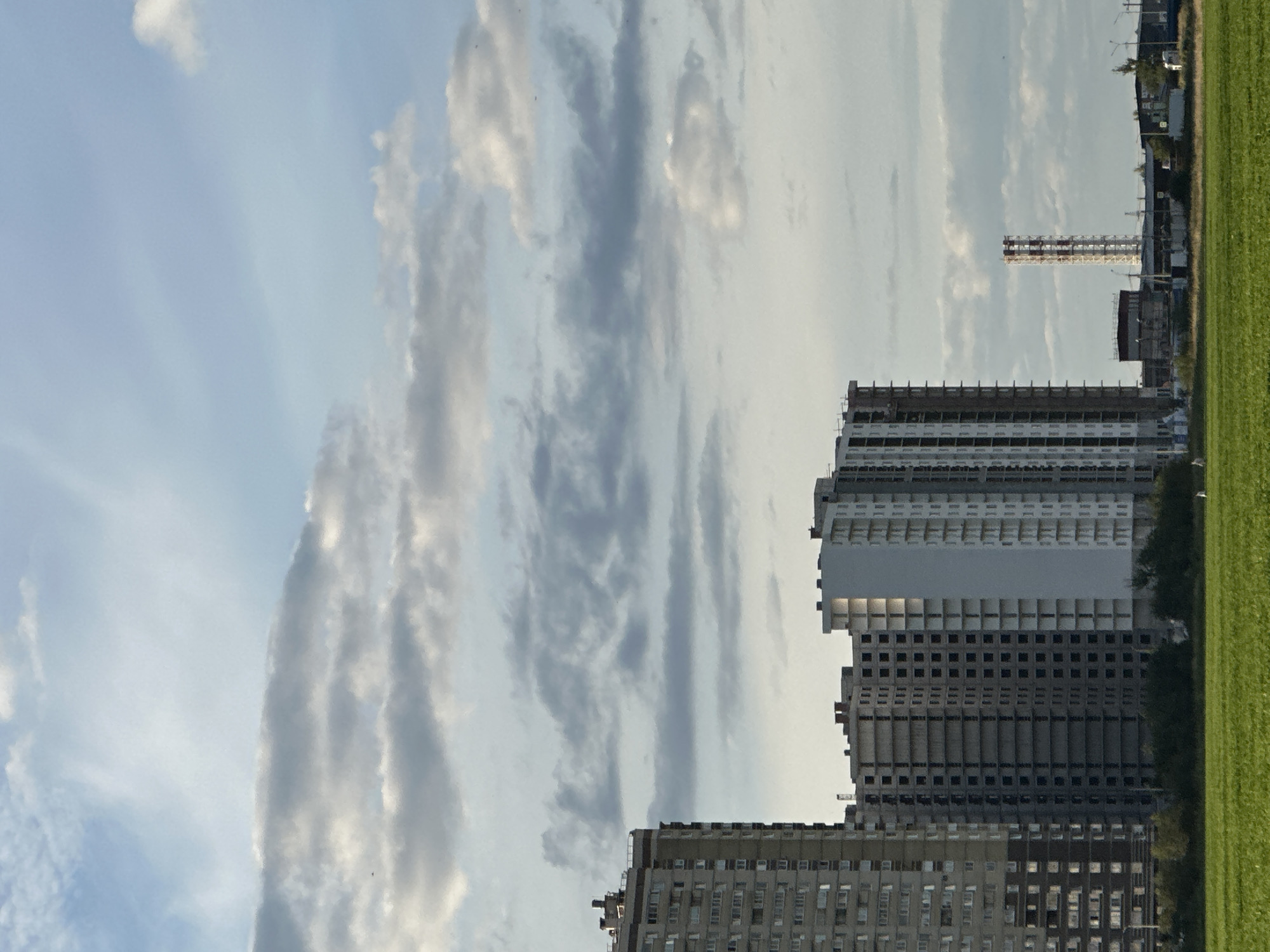 A wide shot captures a partly cloudy sky over a city in Russia. In the foreground, a lush green field stretches across the bottom of the frame. Beyond the field, several multi-story buildings are visible. On the left, a tall, completed apartment building stands, its facade a mix of beige and brown with numerous windows. To its right, a cluster of unfinished apartment buildings rises, their concrete structures exposed. In the distance, a tall industrial chimney with a red and white striped pattern stands against the sky. The overall scene depicts an urban landscape with ongoing construction under a vast, dynamic sky.