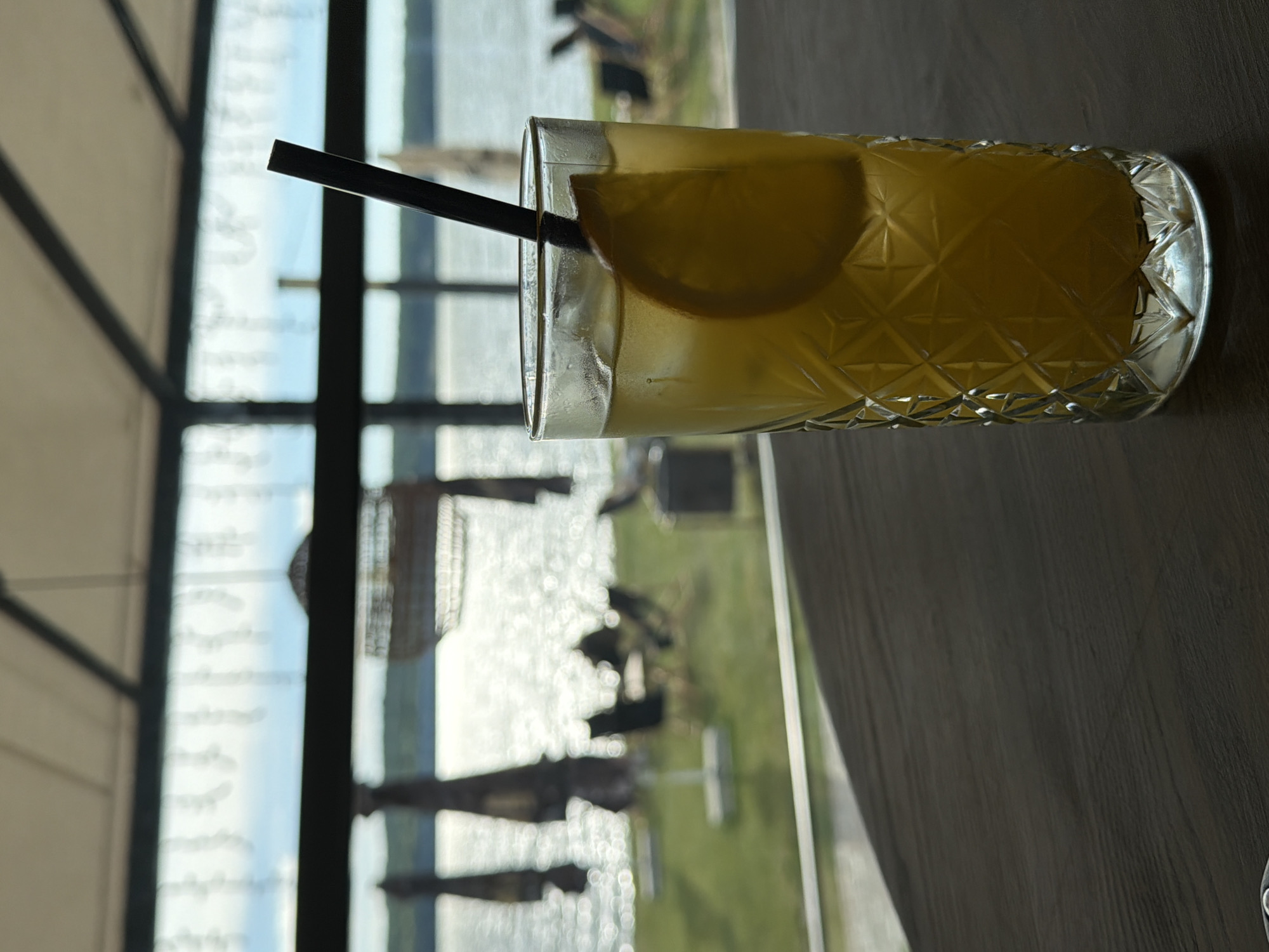 An eye-level, close-up shot shows a tall glass of yellow liquid with ice and an orange slice, with a black straw. The glass has a textured diamond pattern and sits on a dark wooden surface. In the background, a body of water with rippling sunlight reflects off it, and beyond that, a grassy area with several empty chairs and umbrellas. The scene is framed by a ceiling with visible beams and a large window that looks out onto the water. The setting appears to be a cafe or restaurant with an outdoor seating area by a lake. The location is the Ternopil Lake in Ternopil, Ukraine.