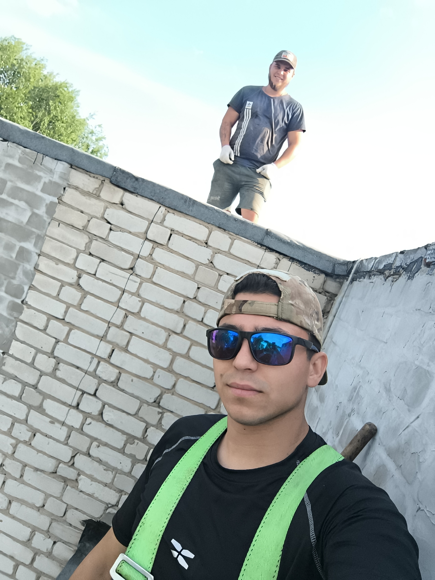 Two men are positioned on a low brick wall structure under a bright sky, with a tree visible in the upper left corner. The closer man, possibly taking a selfie, wears a black shirt with a white logo, green suspender-like straps, a camouflage baseball cap, and sunglasses with blue reflections. Behind him and on top of the wall, a second man with a similar cap, a gray t-shirt, light-colored gloves, and shorts is visible. The wall itself is constructed of light-colored bricks on one side and has a gray, textured surface on the other.