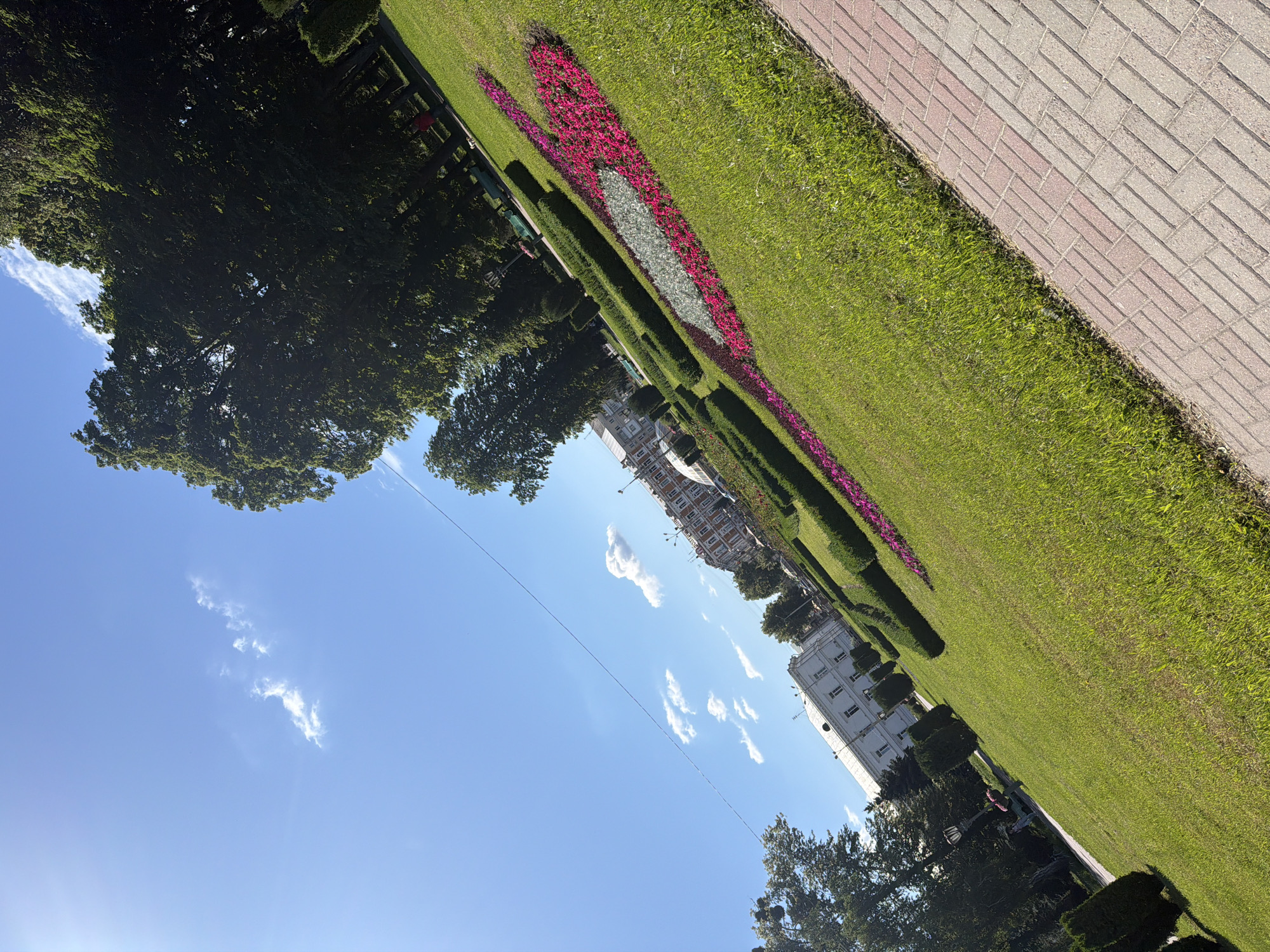 A diagonal, eye-level shot captures a public park on a sunny day. The foreground features a red and gray brick pathway on the right, bordered by a lush green lawn. The lawn is decorated with a flower bed, forming a colorful, elongated shape with pink and white flowers. In the middle ground, manicured green hedges are arranged in geometric patterns. Behind the hedges, buildings with white and beige facades are visible, and tall, dark green trees line the park's edge. The sky above is a clear blue with a few scattered white clouds. A thin black wire stretches across the sky from the upper left to the lower right. The scene is brightly lit by the sun, creating a clean and vibrant atmosphere.