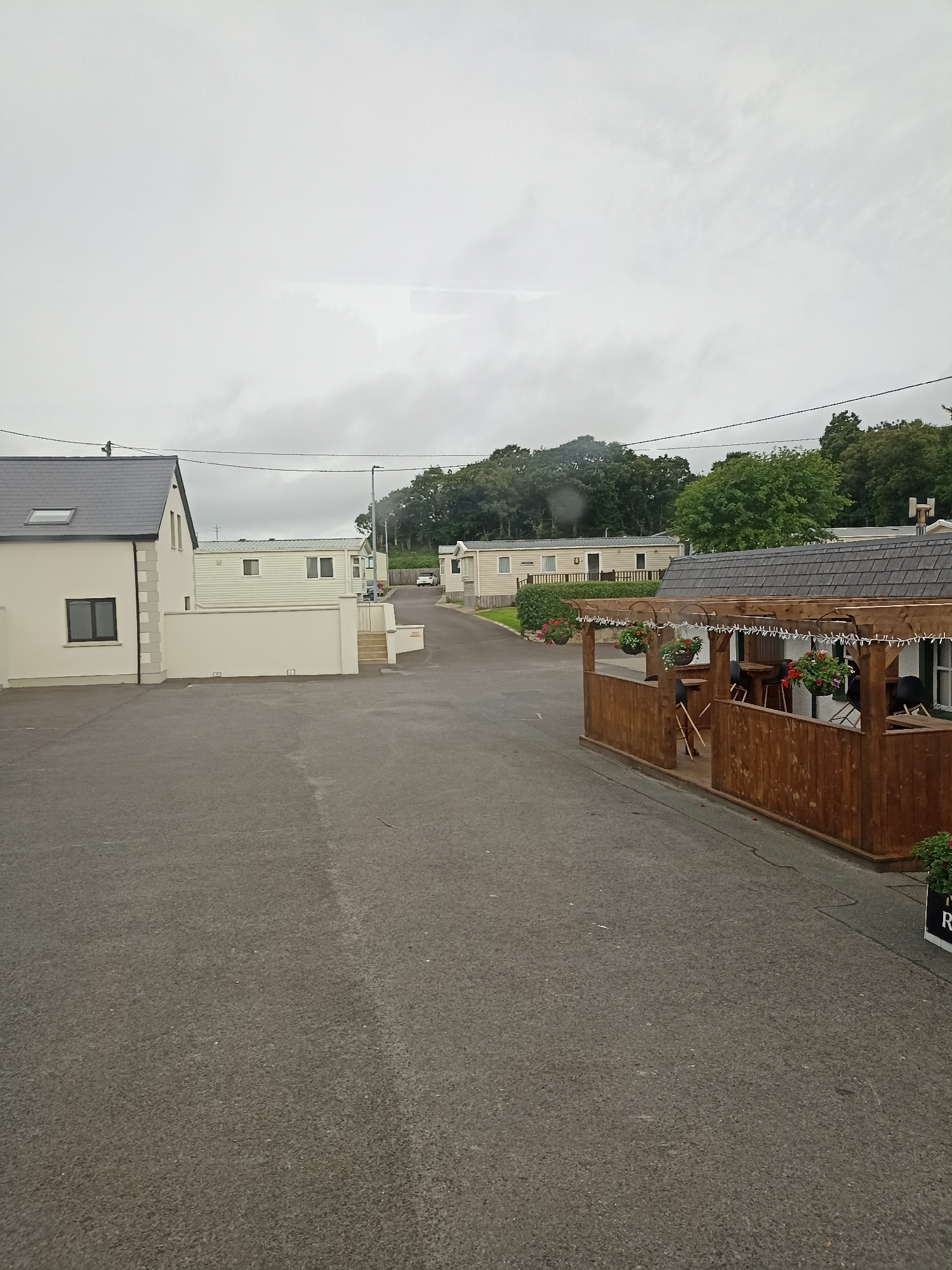 A ground-level, eye-level, full shot captures an overcast day at a caravan park. The foreground is dominated by a dark asphalt parking lot, with a distinct light gray line running diagonally across the pavement. To the right, a wooden patio structure with a dark shingled roof features a railing and hanging flower baskets. A portion of a black and white sign with the letters "R" is visible at the lower right corner. In the mid-ground, a paved road winds through the park, with several static caravans or mobile homes lining the sides. These dwellings are predominantly light-colored with white walls and windows. A single white vehicle is parked on the road near a cluster of trees. A taller, lighter-colored building with a dark gray roof and a skylight is situated on the left side of the frame, with a low white wall in front of it. The background consists of dense green trees covering a gentle slope, under a pale, cloudy sky. The overall impression is of a quiet, possibly residential, outdoor setting.