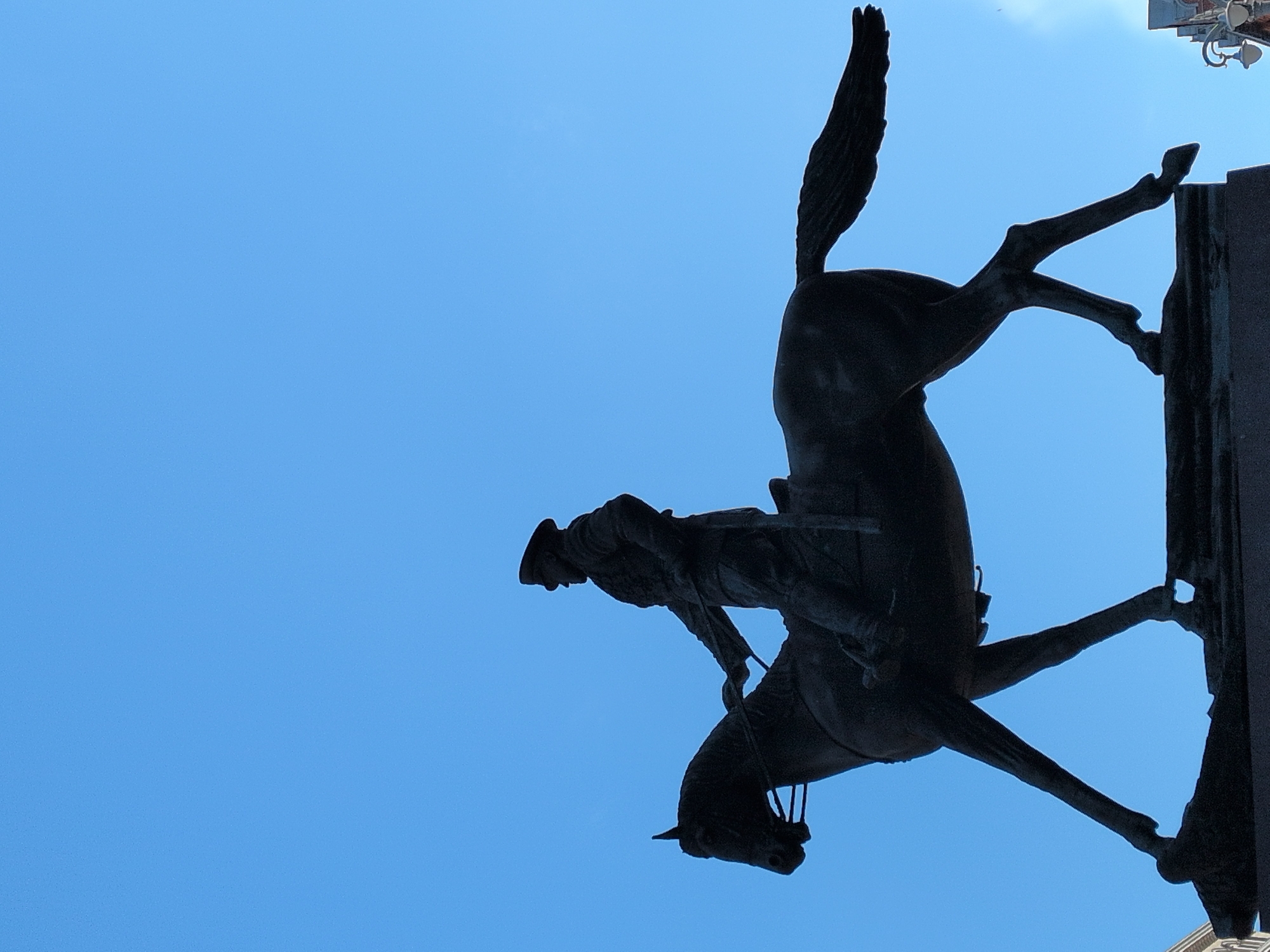 The media file displays an equestrian statue captured from a low angle, looking upwards against a clear, bright blue sky. The entire statue is rendered in deep silhouette, appearing as a dark, almost black form against the vibrant background.
The statue depicts a person, likely a soldier or historical figure, astride a horse that is rearing dynamically on its hind legs with its front legs raised. The rider wears a hat and appears to be holding reins. Due to the photographic perspective or the image's orientation, the statue appears rotated approximately 90 degrees clockwise within the frame.
The background is a uniform, cloudless, and intense blue, indicating bright daylight and clear weather conditions. In the upper right corner of the original image, a small portion of a light-colored structure with some discernible details, possibly part of a building's architecture or an ornamental pedestal, is faintly visible. There is no visible text in the image.