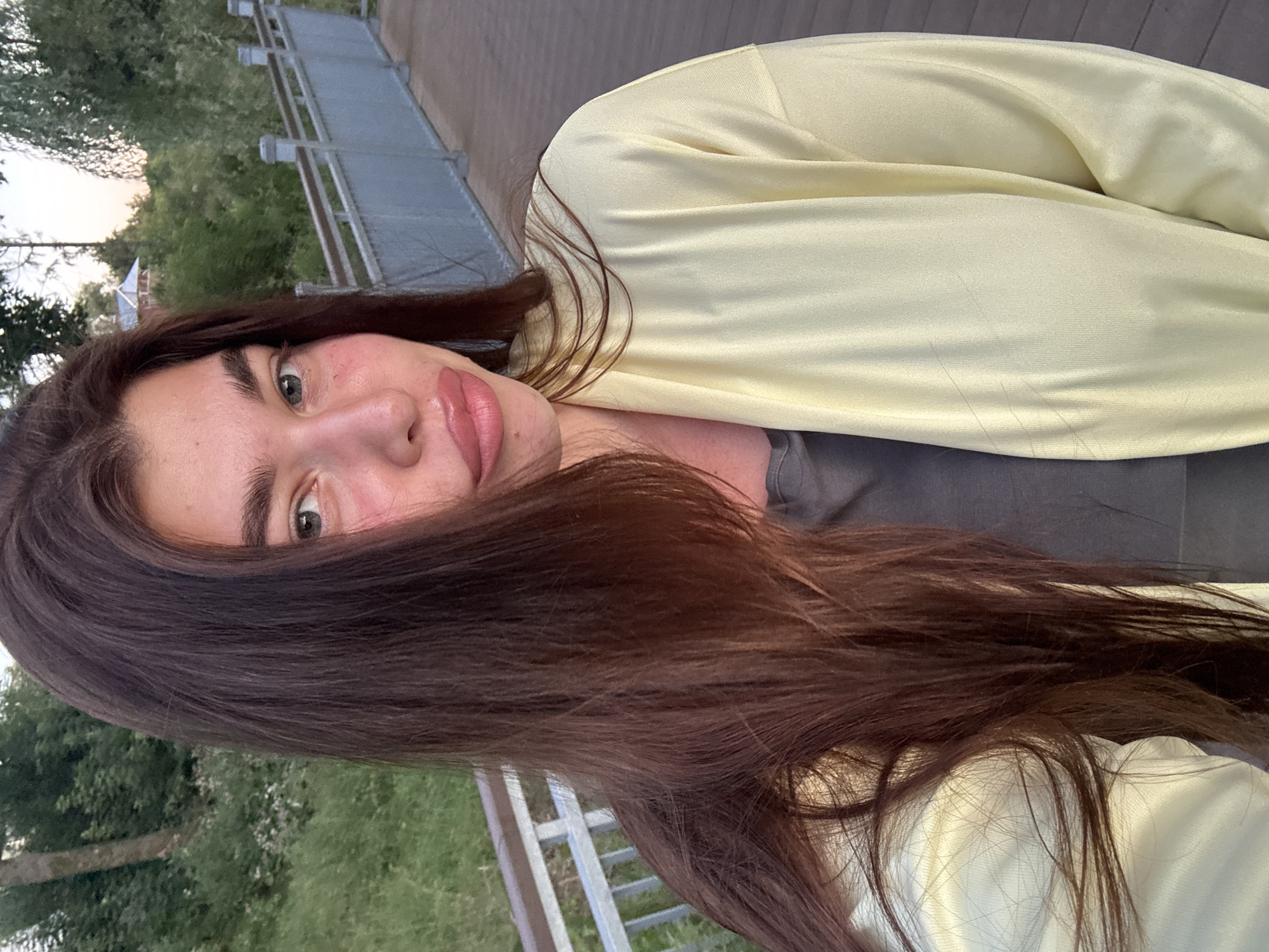 A young woman with long, dark brown hair and light blue eyes is taking a selfie outdoors. She is wearing a gray t-shirt and a pale yellow cardigan. Her lips are slightly parted, and she is looking directly at the camera. Her face appears flushed. Behind her, there is a wooden walkway with a light-colored metal railing. Beyond the railing, there is lush green grass and trees, with a glimpse of a building with a red roof in the distance. The setting appears to be a park or recreational area. The image quality is clear and well-lit, suggesting it was taken during daylight hours. The location is identified as Klin, Russia.