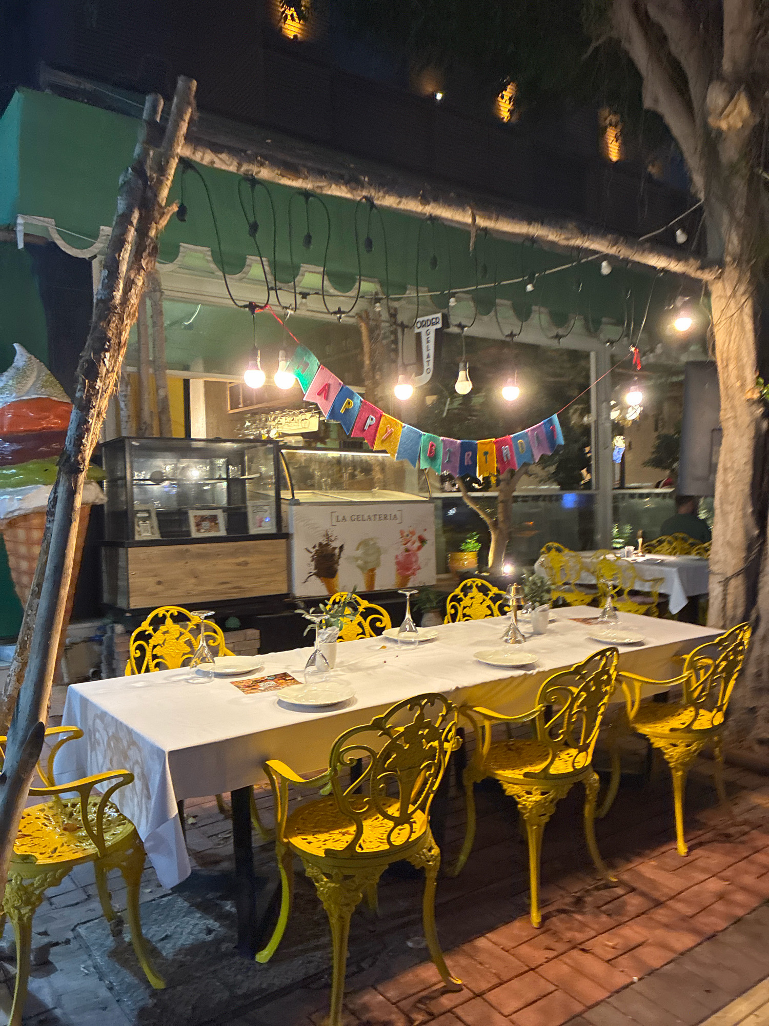 An outdoor seating area for a gelato shop in Alanya, Turkey, is set up for a birthday celebration at night. A long, white-clothed table is set with plates and glasses, surrounded by ornate yellow chairs. A string of colorful "HAPPY BIRTHDAY" flags hangs overhead, along with various lightbulbs that illuminate the scene. In the background, a glass-fronted display case shows various ice cream flavors, and beyond that, a large window looks into the dimly lit interior of the shop. A large, three-scoop ice cream cone decoration stands to the left of the frame, and a tree with string lights adds to the festive ambiance. The ground is paved with red bricks.