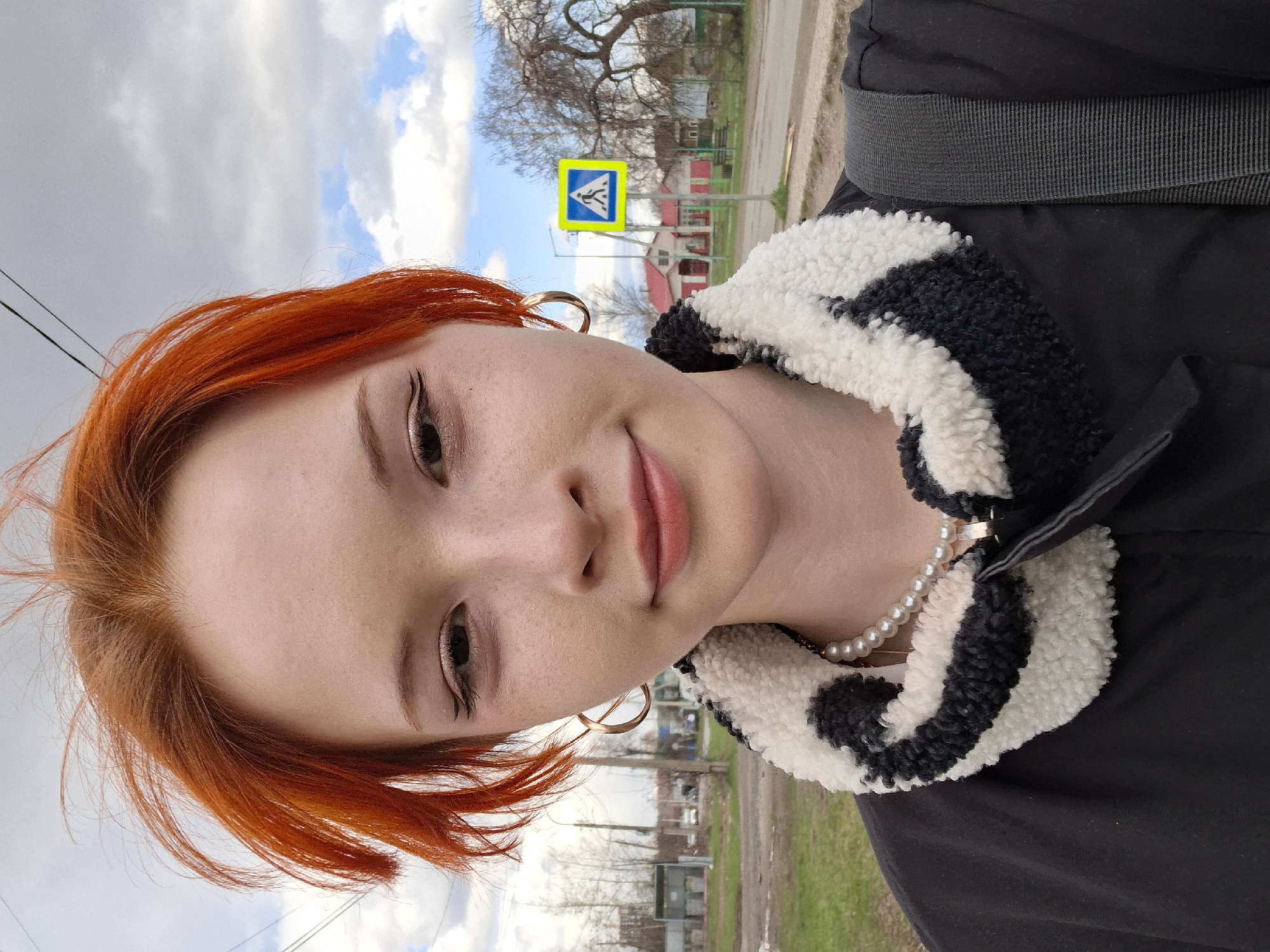 A selfie-style portrait features a young person with vibrant red-orange hair, light skin, and a subtle, gentle smile. They are wearing gold-toned hoop earrings and a pearl necklace, complementing a black jacket with a distinct black and white striped, fluffy collar. A black strap, possibly from a backpack, is visible over one shoulder.
The outdoor setting indicates daytime under a partly cloudy sky with patches of blue. In the background, a prominent yellow square road sign with a blue border displays a white pedestrian crossing symbol. Beyond the sign, several bare trees suggest a cooler season, such as late autumn or early spring. Distant buildings with red roofs and light-colored walls, along with a road or path and grassy areas, complete the scene. The overall impression is a casual moment captured in an urban or suburban outdoor environment, consistent with the location context of Chaikovsky, Russia. The person's expression is relaxed and pleasant.