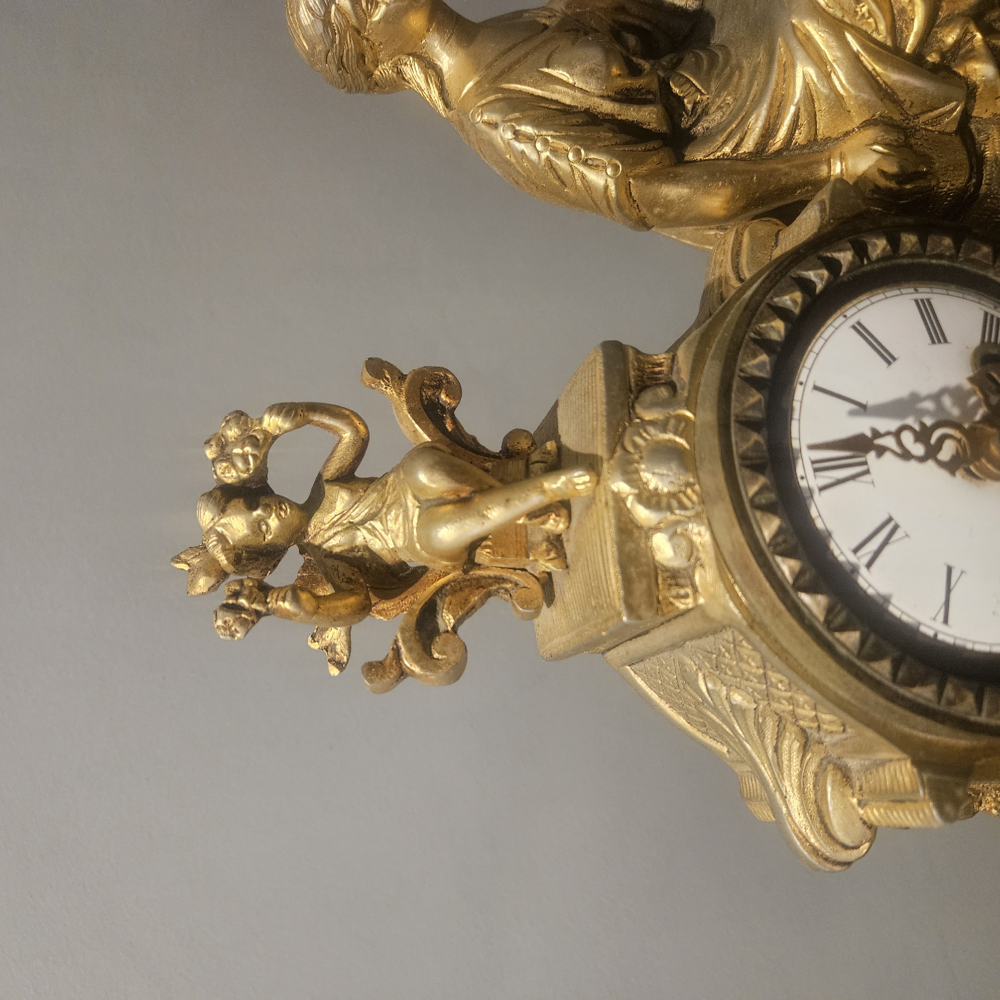 The image shows a close-up, eye-level shot of a decorative golden clock. The clock is adorned with two classical figures: a cherubic figure with wings and holding flowers sits on a pedestal on the left, and a draped female figure sits on the right. The clock face, with Roman numerals, is partially visible at the bottom. The background is a plain, neutral gray wall. The image is taken in the city of Almadrones, Spain, likely indoors. There are no people or animals present. The lighting appears to be soft and diffused, highlighting the metallic sheen of the clock.