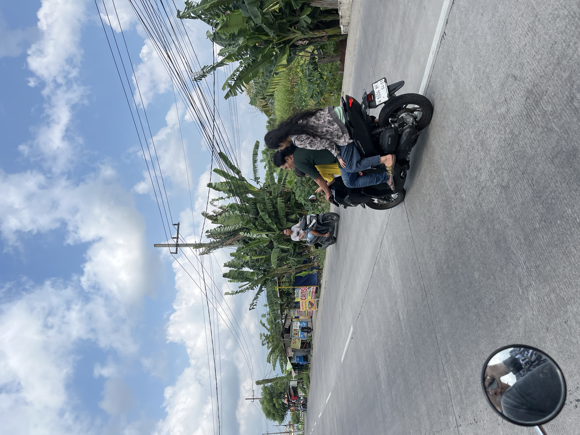 This image depicts a street scene in Kepatihan, Indonesia. In the foreground, a dark-colored motorcycle is traveling on a paved road. The motorcycle carries three occupants: a male driver wearing a green shirt and jeans, a female passenger behind him in a patterned jacket and jeans, and a young person in a yellow shirt between them. The motorcycle has a license plate that reads "H 6054 WH 04-21". The road has white lane markings. To the right of the road are numerous banana trees and other lush green vegetation. In the distance, there are several small buildings, possibly shops or stalls, lining the road, with signs visible. Utility poles and overhead electrical wires are present. The sky is blue with scattered white clouds. In the lower left corner, a motorcycle's rearview mirror reflects the photographer, who appears to be holding a phone. Another motorcyclist in a light-colored shirt and blue shorts is visible further down the road on the right, also heading in the same direction. Several other motorcycles are parked or in motion in the background near the buildings.