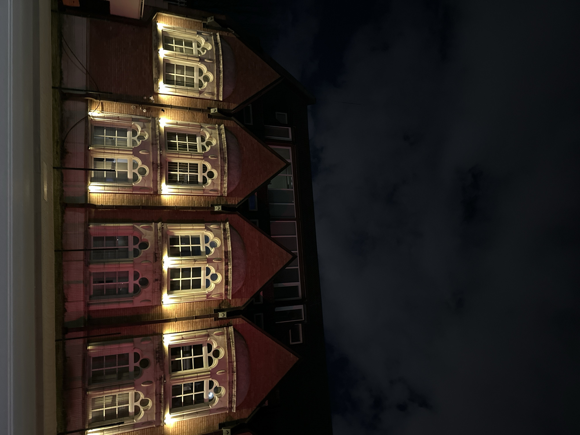 At night, a brick building with ornate, lit windows stands against a dark sky. The building has a distinct architectural style with gabled roofs over each window section. The windows themselves are divided into multiple panes. The location is not evident from the image alone, thus it is not possible to provide location information for the city of Sofia, Bulgaria.
