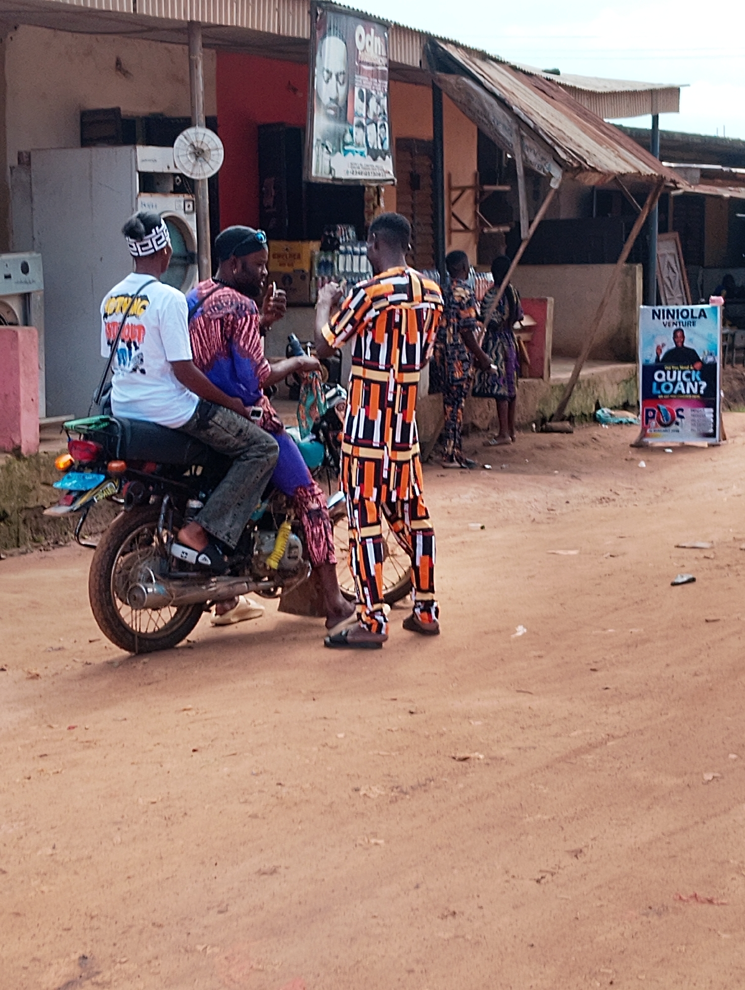 In Orudu-Olokan, Nigeria, three individuals are gathered near a motorcycle parked on a dirt road. One person, wearing a white t-shirt with black lettering and a white patterned headband, sits at the rear of the motorcycle. Another individual, dressed in a patterned blue and red garment, sits behind the driver, who is wearing a colorful orange, black, and white patterned outfit and black sunglasses. The driver appears to be speaking or gesturing with his left hand, holding a small object.
Behind the individuals, a weathered building with a corrugated metal roof stands. A sign advertising "NINIOLA VENTURE" with the tagline "QUICK LOAN?" and the logo "POS" is prominently displayed. The environment is dusty and appears to be an outdoor market or roadside commercial area, with other buildings and signs visible in the background. The overall scene suggests a casual interaction or transaction taking place in a bustling, informal setting.