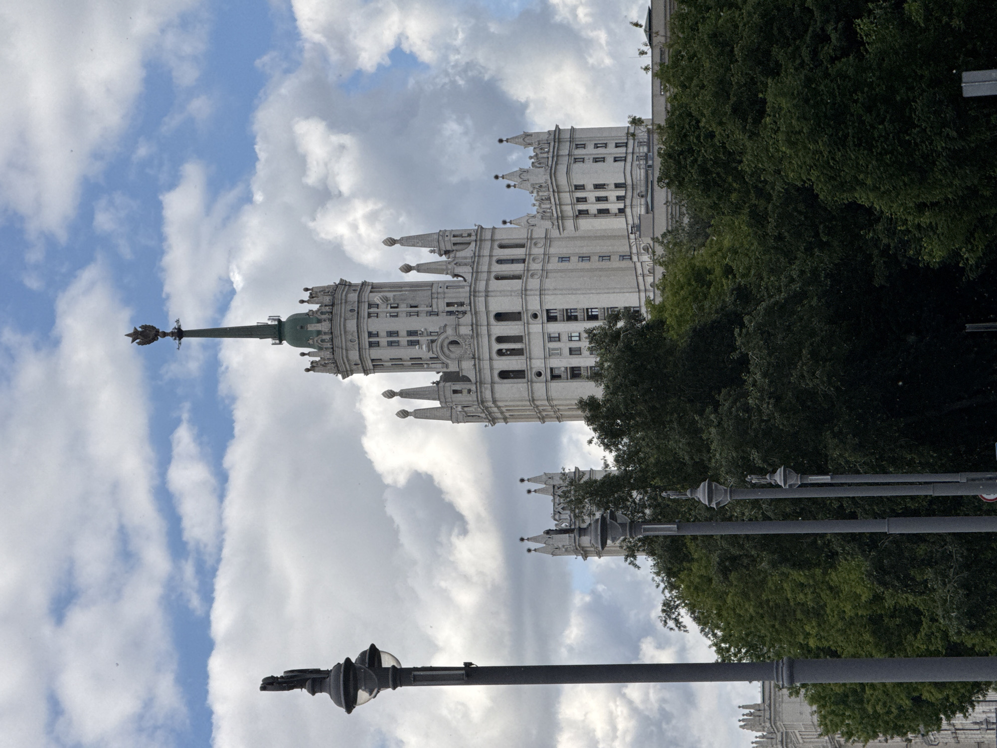 The image shows the Kotelnicheskaya Embankment Building in Moscow, Russia. The building is a Stalinist skyscraper with a tall, tiered structure and a spire topped with a red star. The sky is partly cloudy. The scene is viewed from ground level. Streetlights are visible in the foreground, along with foliage from nearby trees.