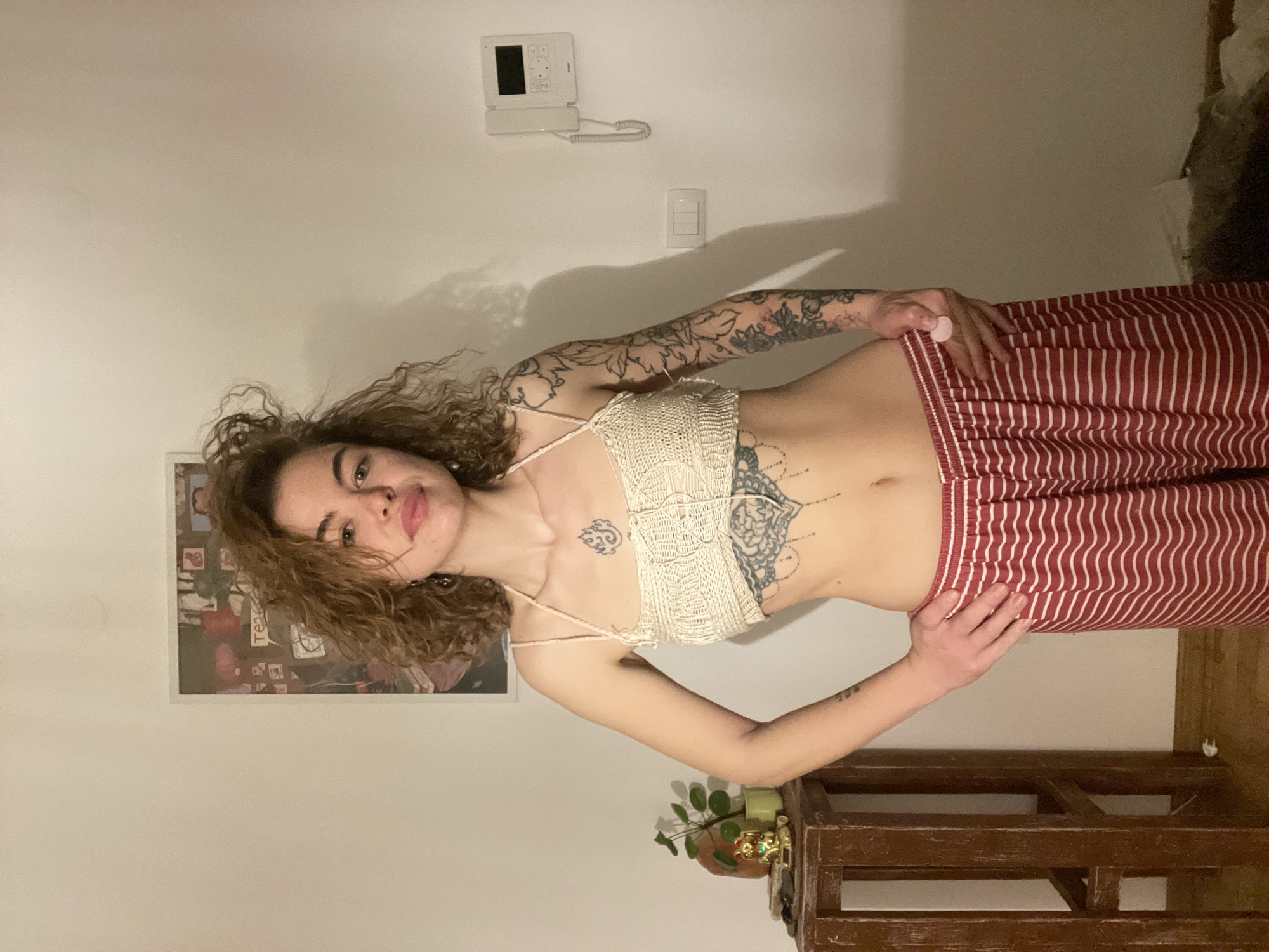 A woman with curly brown hair is standing indoors, posing for a photo. She is wearing a white crochet crop top and red and white striped pants. She has tattoos on her arms, chest, and abdomen. Her left hand rests on her hip, and her right hand is near the waistband of her pants. The background includes a white wall with an intercom system, a light switch, a framed picture, and a dark brown wooden structure (possibly a shelf or table). A potted plant sits on the wooden structure. The location is likely a residence, but there is no evidence that the image was taken in Boreti, Montenegro.
