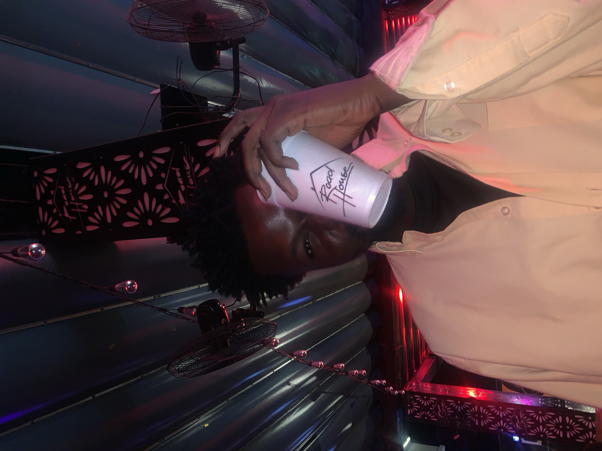 A person is holding a white cup in front of their face in a dimly lit, indoor setting. The cup has a logo that reads "Road House" written in black cursive. The person is wearing a light-colored collared shirt. In the background, there are ceiling fans, string lights, and a patterned decorative panel with red lighting. The environment appears to be a bar or entertainment venue. The location is not specified in the image or crops.