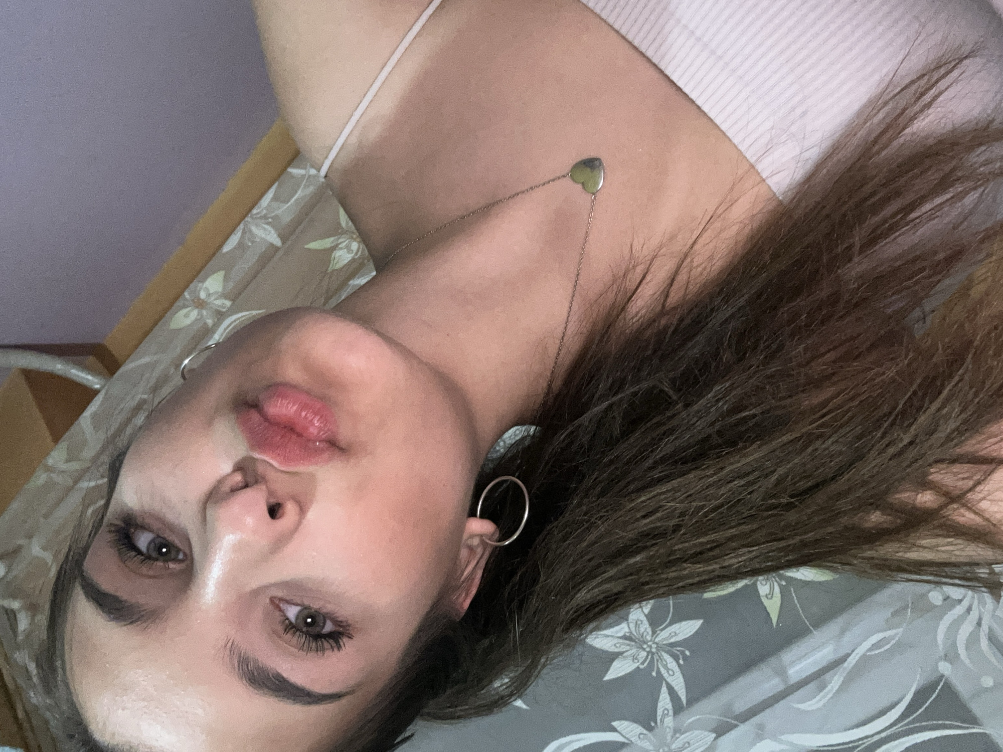 The image shows a close-up, head-and-shoulders portrait of a young woman with long, dark brown hair. She has light brown eyes, defined eyebrows, and is wearing makeup, including mascara and lip gloss. She is making a kissy face, with her lips slightly parted. She is wearing a white tank top or camisole and a silver necklace with a heart-shaped pendant. She also has a silver hoop earring in her left ear. The background appears to be an indoor setting, possibly a bedroom, with a patterned curtain or wallpaper visible behind her. There are no visible landmarks that would indicate a specific location in Shumen, Bulgaria.
