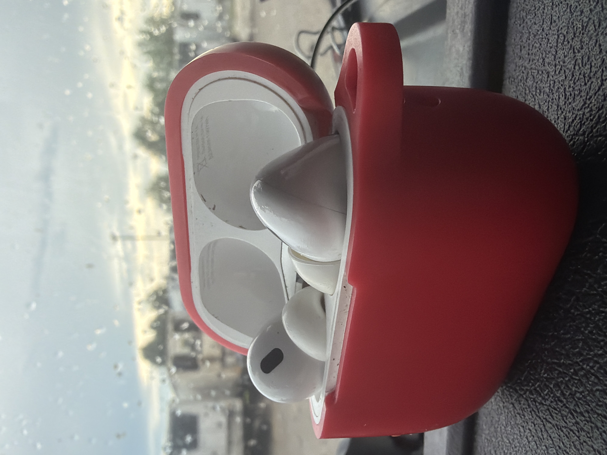 The image shows a close-up, eye-level view of an open red charging case for wireless earbuds. The earbuds themselves are white and are partially visible within the case. The charging case is positioned on a dark, textured surface, possibly a dashboard or a car interior. In the background, a window shows a blurry outdoor scene with trees, vehicles, and a cloudy sky with visible raindrops on the glass. The overall impression is a still life of personal electronic devices set against a backdrop suggesting travel or an outdoor environment in Murfreesboro, United States of America.