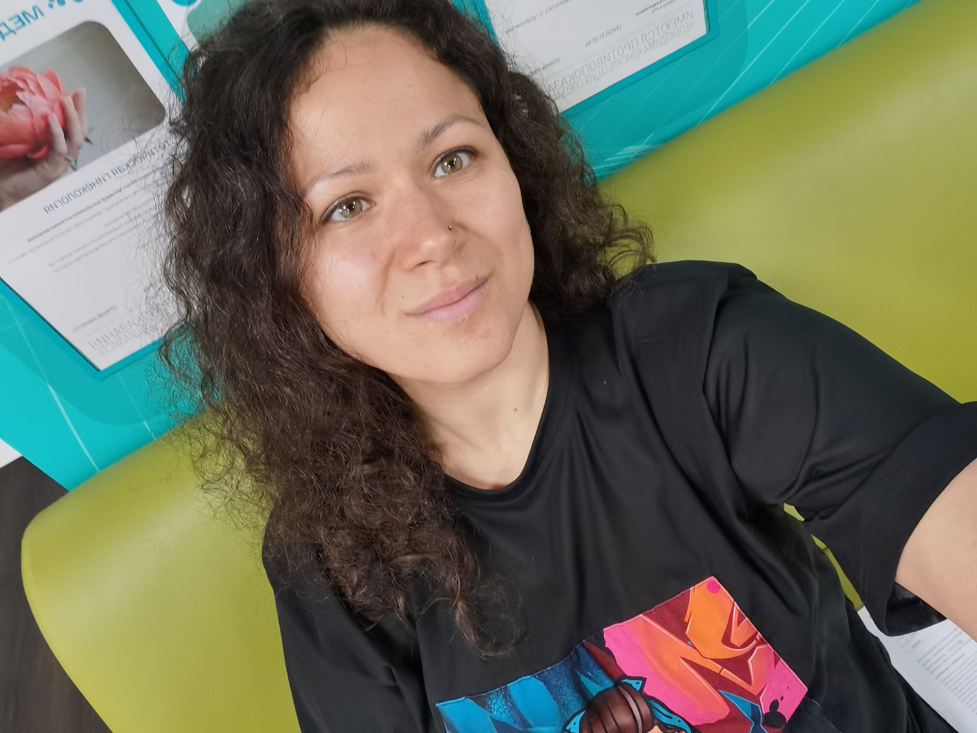 A young adult female is depicted in a medium shot, seated or leaning against a lime-green upholstered surface, presumably a bench or sofa. She has dark, curly hair and light brown or hazel eyes, with a small stud piercing on her left nostril. Her expression is a subtle smile, and she appears to be looking slightly upwards and to the viewer's right. She is wearing a black, short-sleeved t-shirt featuring a colorful, graffiti-style graphic design on the chest, incorporating shades of pink, orange, and blue. The background to her left (viewer's right) reveals a turquoise-colored wall or panel with horizontal white stripes. Partially visible behind her left shoulder is a white document or poster with text rendered in Cyrillic script, which appears to be mirrored, along with an image of a hand holding a pink flower. The presence of Cyrillic script is consistent with a location in Russia, specifically, as provided, the city of Izhevsk.
