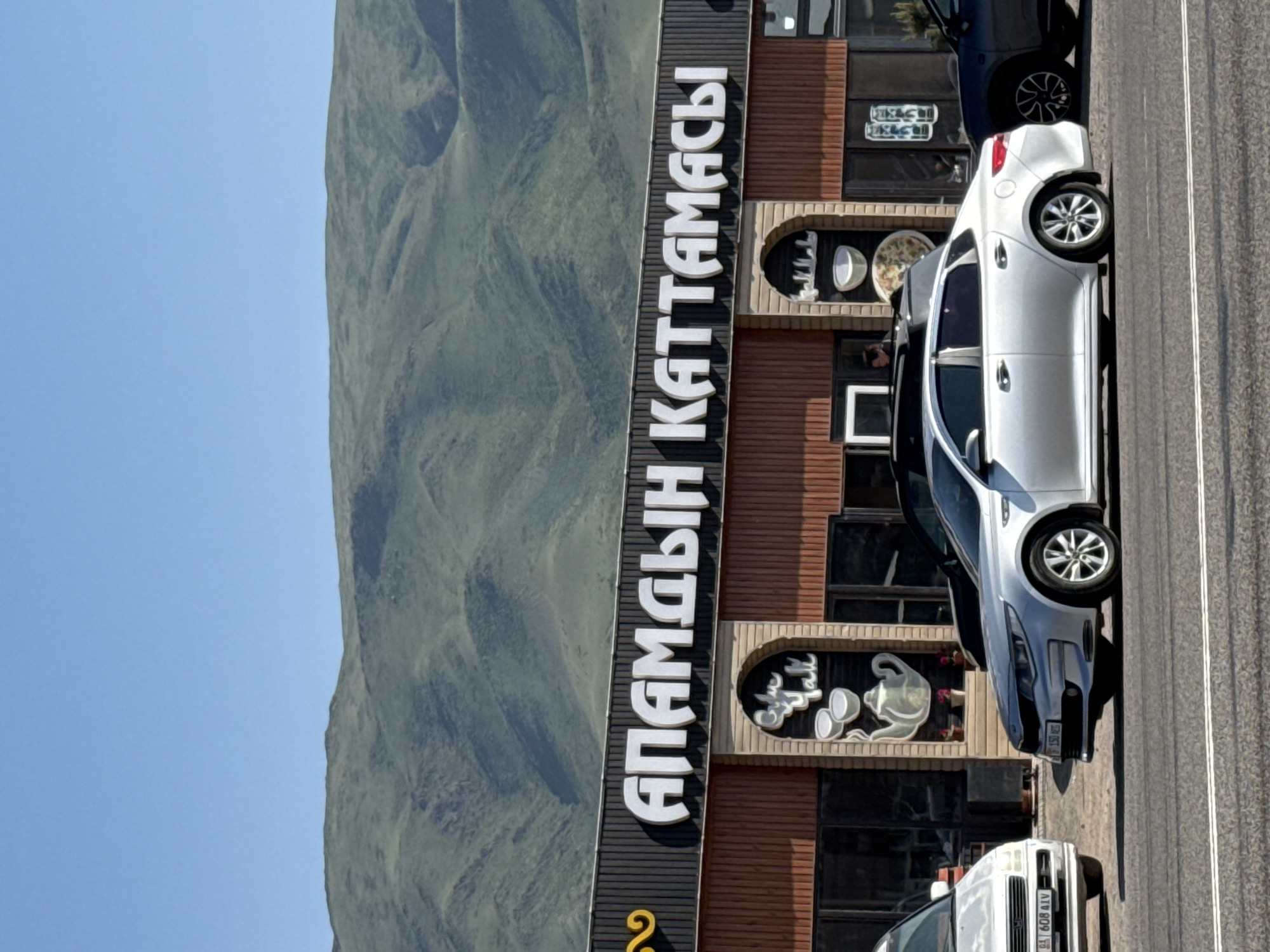 The image depicts a roadside scene in Sasyk-Bulak, Kyrgyzstan. A building with a large vertical sign reading "АПАМДЫН КАТТАМАСЫ" stands alongside a road. Cars are parked in front of the building. A mountain range is visible in the background. The sky is clear and blue.