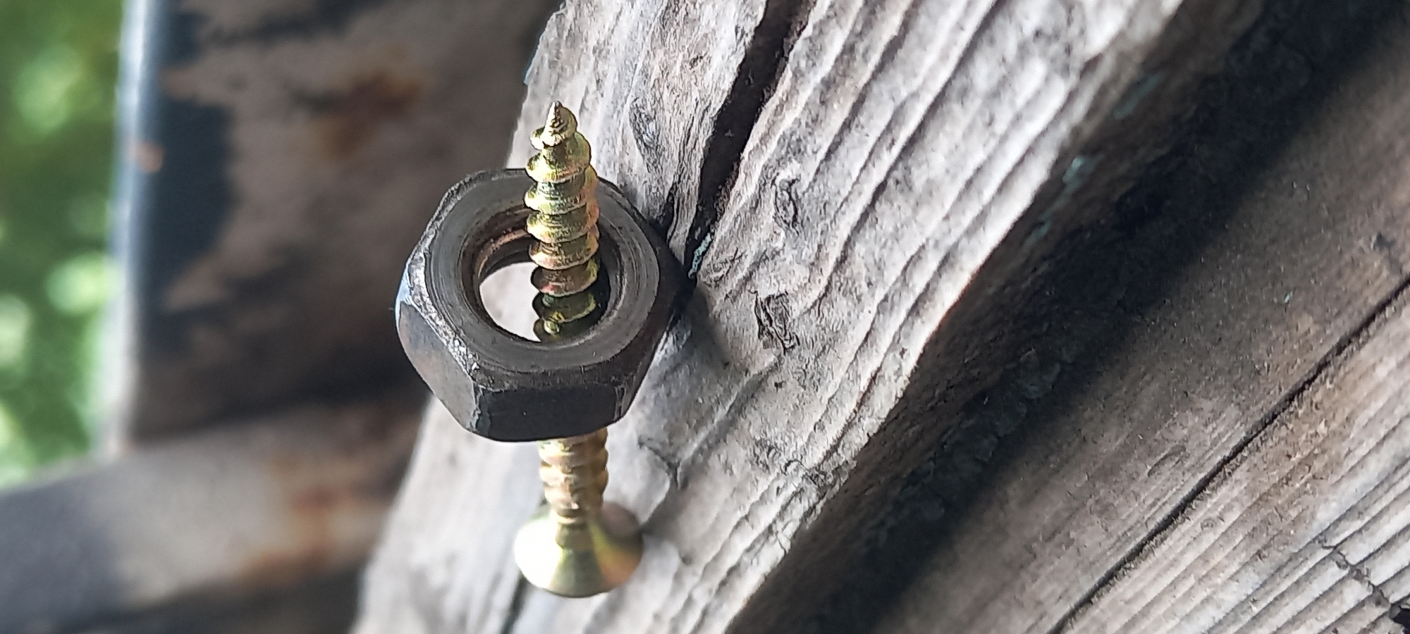 A macro photograph shows a single metal nut with a screw threaded through its center. The nut is hexagonal and dark gray, while the screw is golden yellow with prominent threads. The screw is partially inserted into the nut, with its pointed end visible. Both objects are resting on a weathered, rough wooden surface that is likely an outdoor structure. The background is blurred, showing hints of green foliage and possibly other wooden elements. The image is sharpest on the screw and nut, drawing attention to their texture and detail. The scene is devoid of any human presence or activity and appears to be located in a natural or semi-natural outdoor environment. There are no identifiable landmarks that would place this image specifically in Kryvyi Rih, Ukraine.