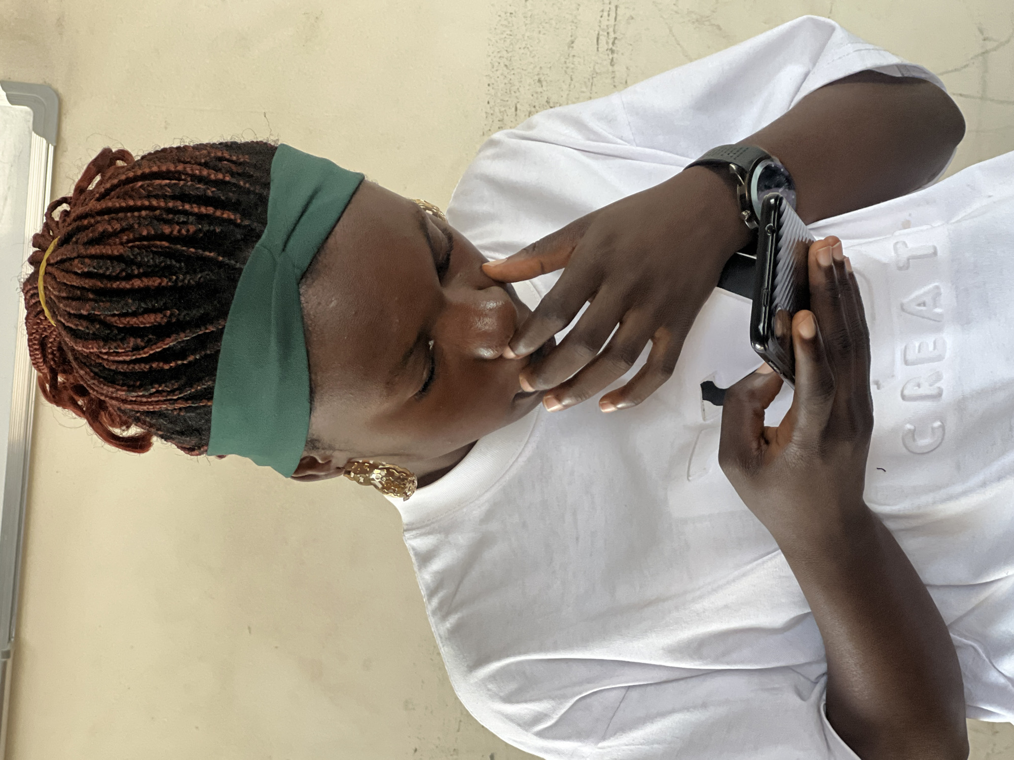 A close-up, medium shot shows a young Black woman from the shoulders up, looking down at a smartphone in her hands. She has braided hair, reddish-brown in color, pulled up and secured with a yellow band, and wears a green knotted headband. Large, gold, hammered-style earrings dangle from her ears. She is wearing a white t-shirt with a raised, embossed design that appears to spell "CREATE" across the chest. Her right hand is near her mouth, with her fingers touching her nose. On her left wrist, she wears a black watch with a textured band. The background is a plain, light beige wall. The image is not suggestive of any specific location in Jos, Nigeria, beyond a generic indoor or outdoor setting with a wall.