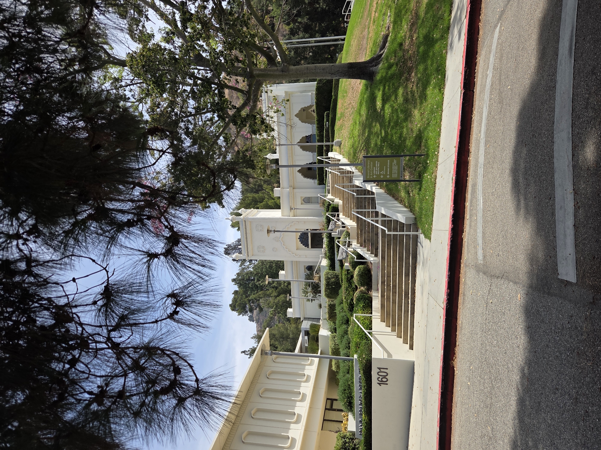 This is a full, slightly downward-angled, outdoor shot of the Brand Library & Art Center in Glendale, California, on a sunny day. The building is white with classical-inspired architecture, featuring arched windows and decorative turrets. A long, wide staircase leads up to the main entrance of the art center, with manicured hedges and bushes lining the sides of the stairs. To the left of the staircase, a smaller, modern white building with rectangular window-like indentations is visible, with "1601" printed on its side. Several trees, including a large one with exposed roots to the right of the building, and pine trees in the foreground casting shadows, are present. A street with a red curb is in the foreground, with a signpost to the right of the stairs providing directions to various parts of the art center. No people are visible in the frame.